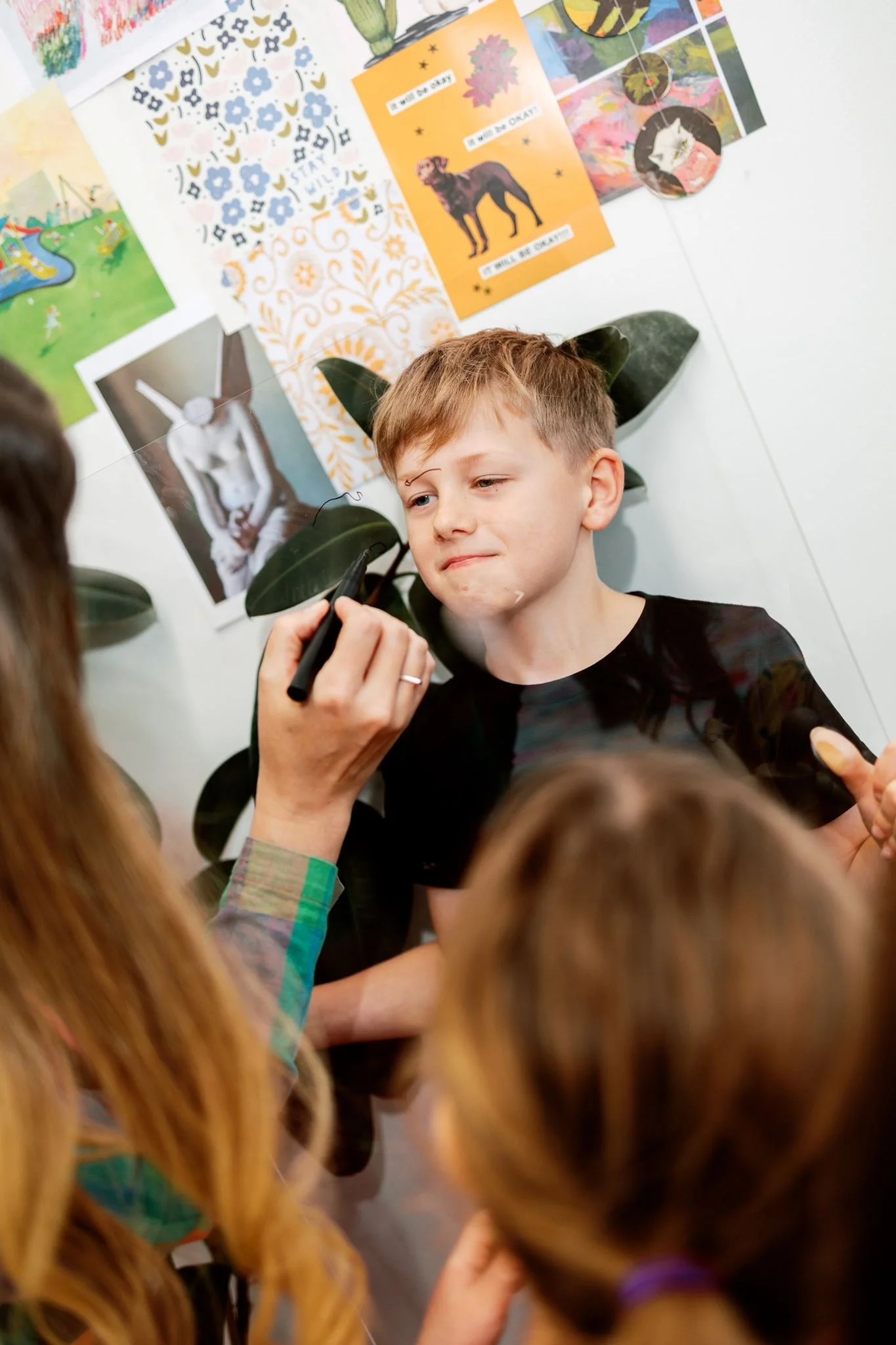 A young boy with short hair and a black shirt is getting a face paint or makeup application by a woman with long hair who has a bracelet on her wrist. The background features a white wall decorated with colorful artwork, including posters, drawings, and a picture of a dog on an orange background.