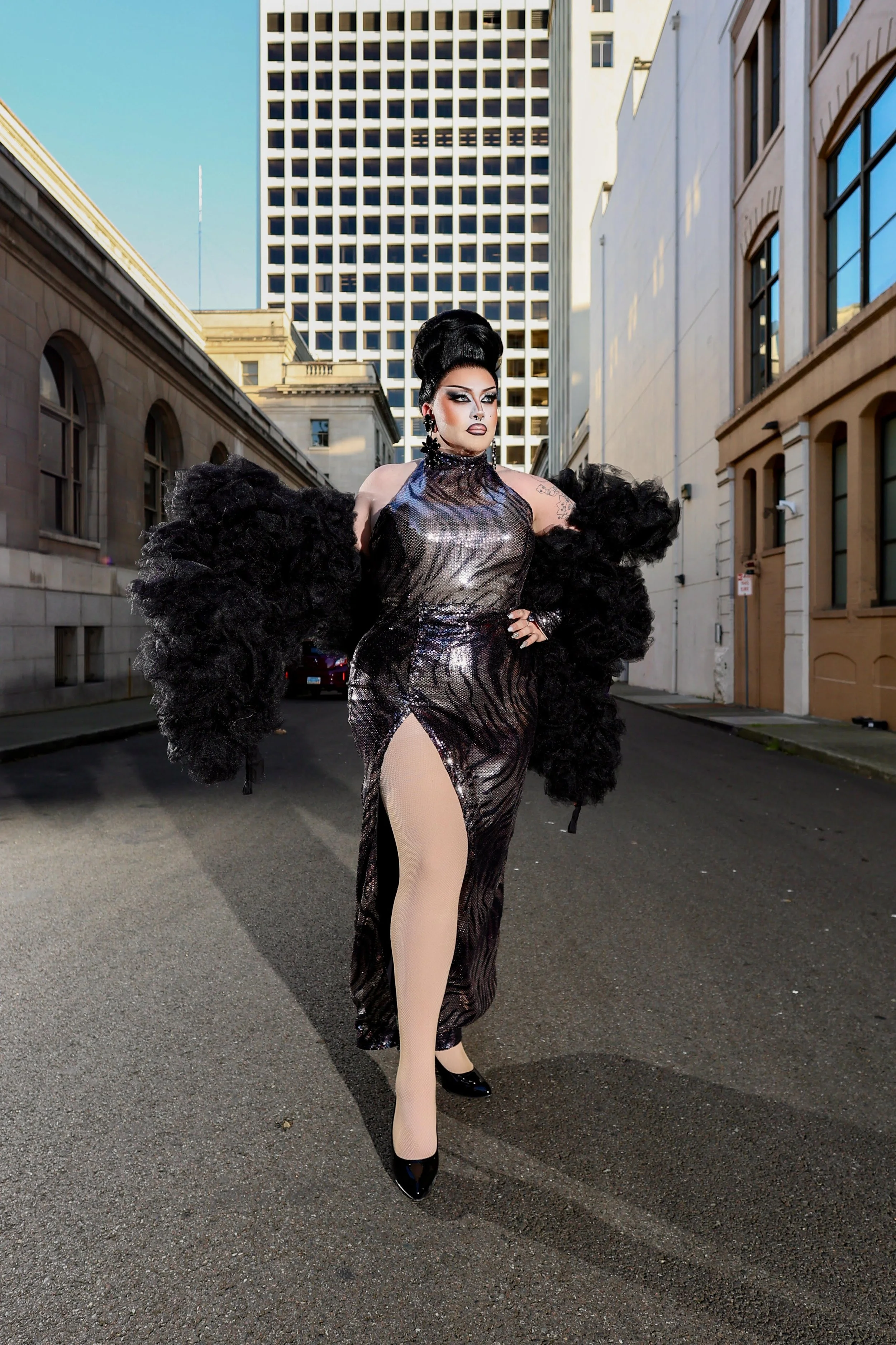 A person with exaggerated makeup and a high, styled black hairstyle stands in the middle of a city street. They are wearing a shiny, black, form-fitting dress with a thigh-high slit, beige fishnet stockings, and black high heels. Large black feather-like accessories are draped over their shoulders.