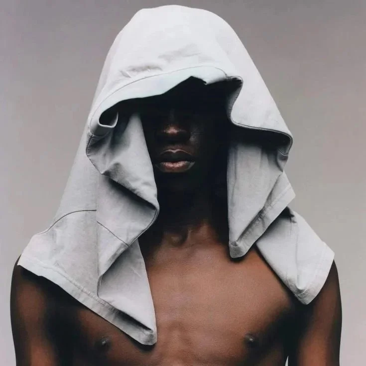 A person with dark skin, shirtless, has a gray hoodie draped over their head, obscuring their face against a plain gray background.
