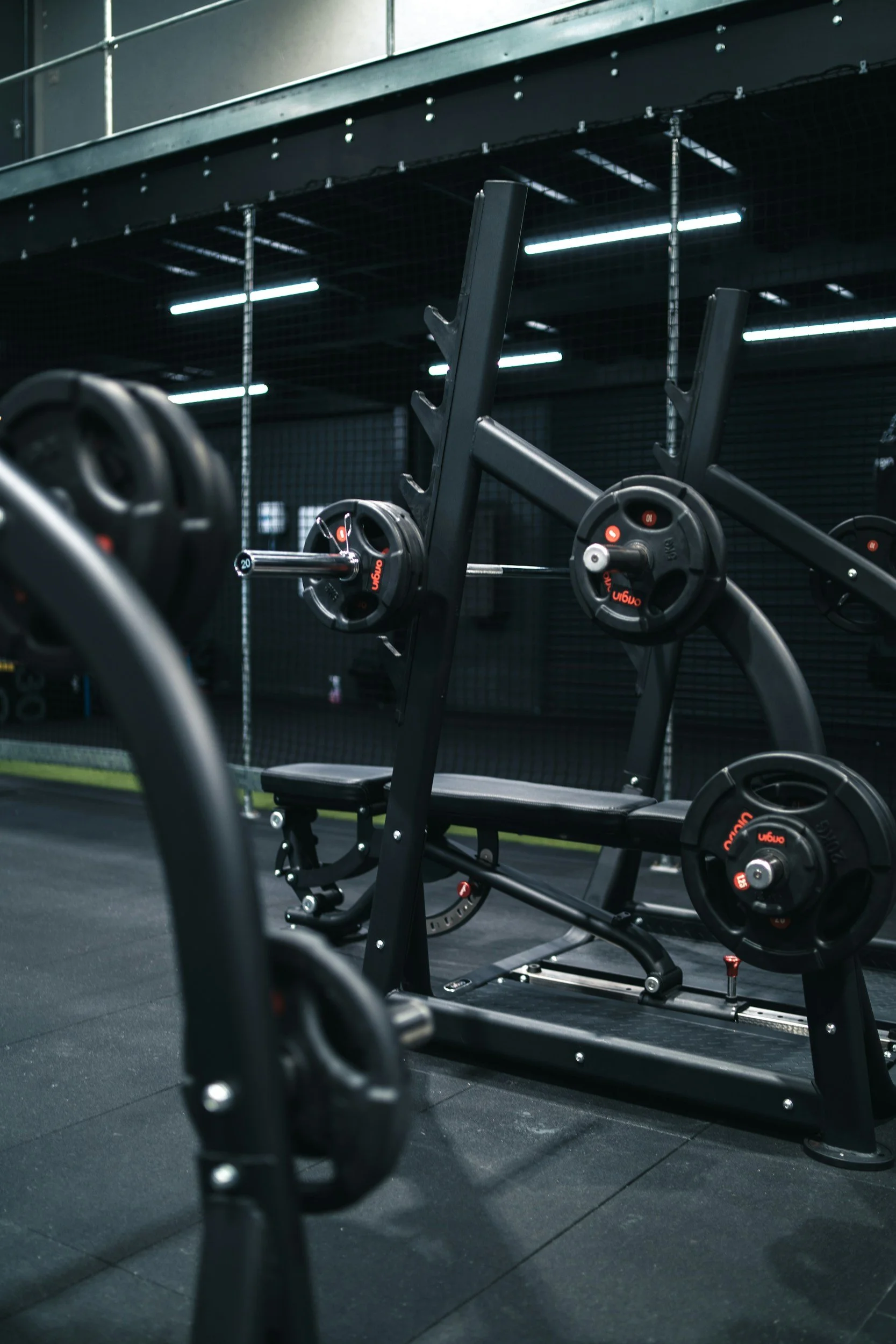 Inside a gym, black workout equipment including weight plates and a bench is visible, with a black grid wall and fluorescent lights above.