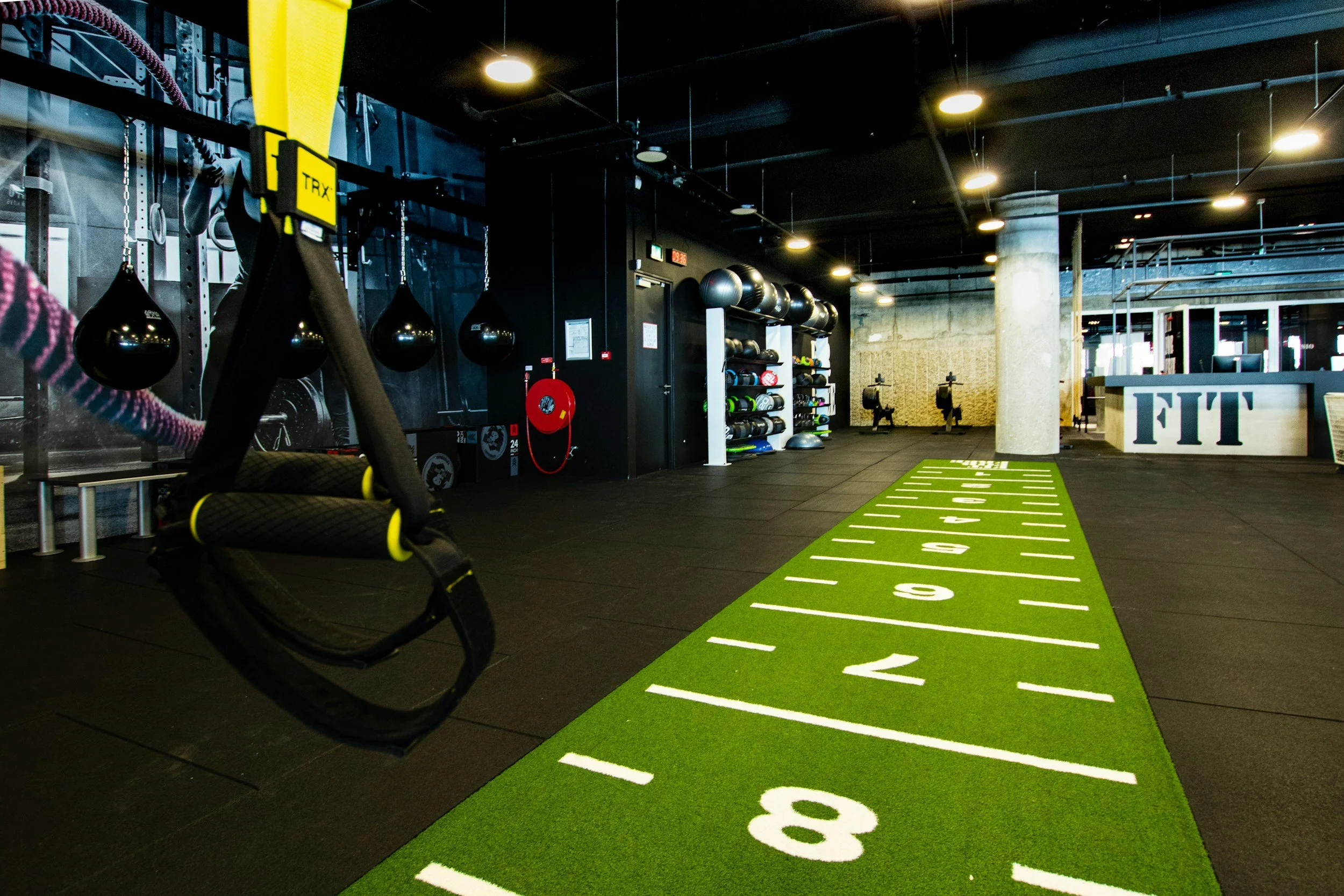 Interior of a modern gym featuring fitness equipment, boxing bags, a green running track marked with numbers, and shelves with exercise gear.