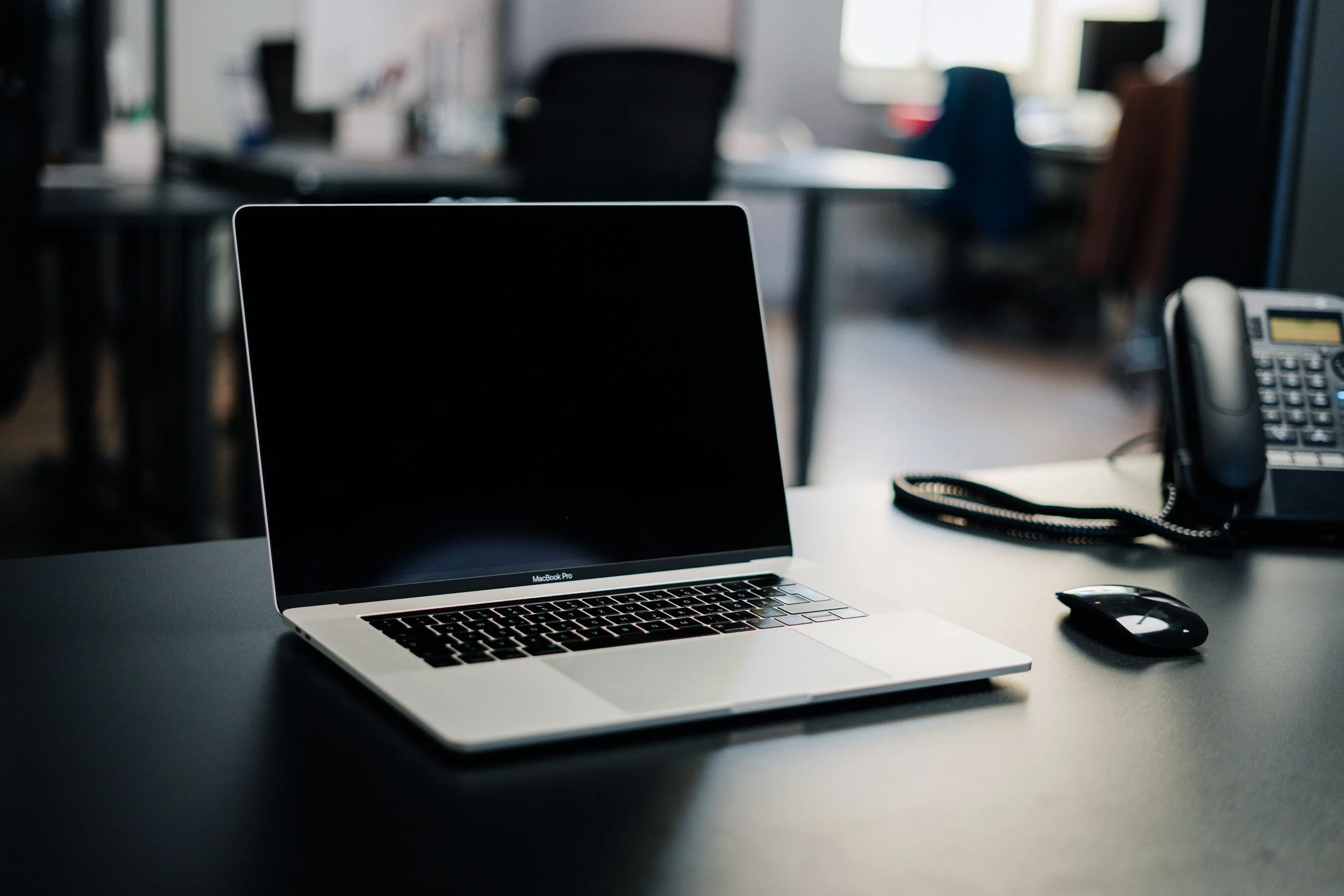 An open MacBook Pro laptop on a white office desk, with a computer mouse nearby. In the background, there are office chairs, desks, and a desktop phone on the right side of the image.
