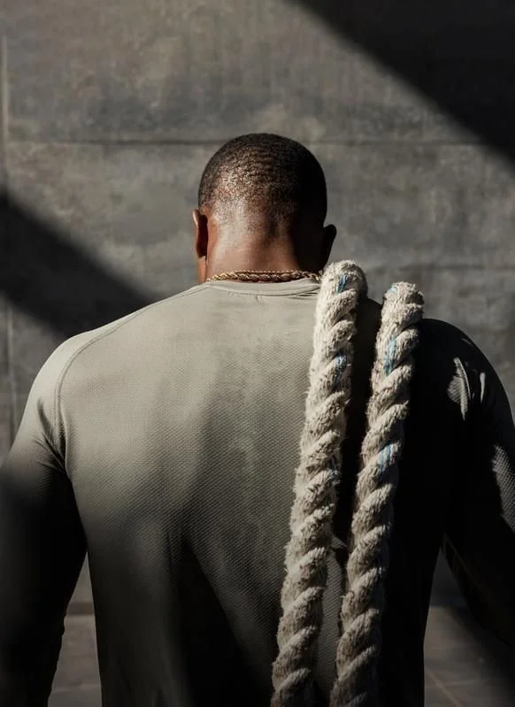 Back view of a man wearing a gray athletic shirt, with a thick rope draped over his shoulder, in a dimly lit setting with gray concrete wall background.