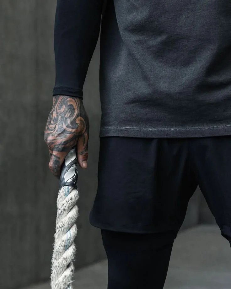 A close-up of a person's torso and hand holding a thick, twisted rope. The person is wearing a dark long-sleeve shirt and dark shorts or athletic wear, with visible tattoos on their hand and arm.
