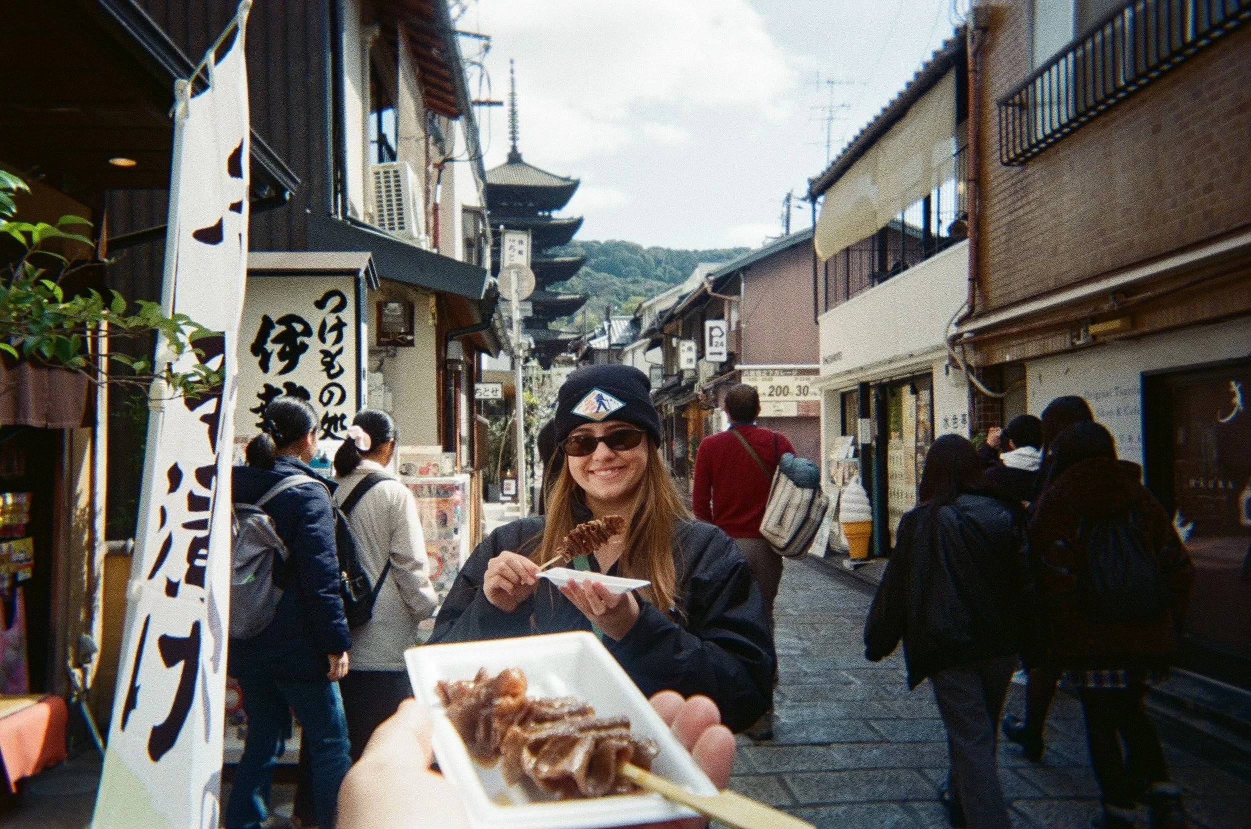 A woman in a black jacket and sunglasses smiling while holding a plate of food, with a busy street and traditional Japanese buildings in the background.