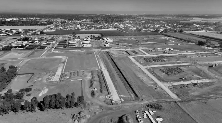 Aerial view of an undeveloped area with construction underway, showing dirt roads, trenches, and partially built structures, with a town and farmland in the background.