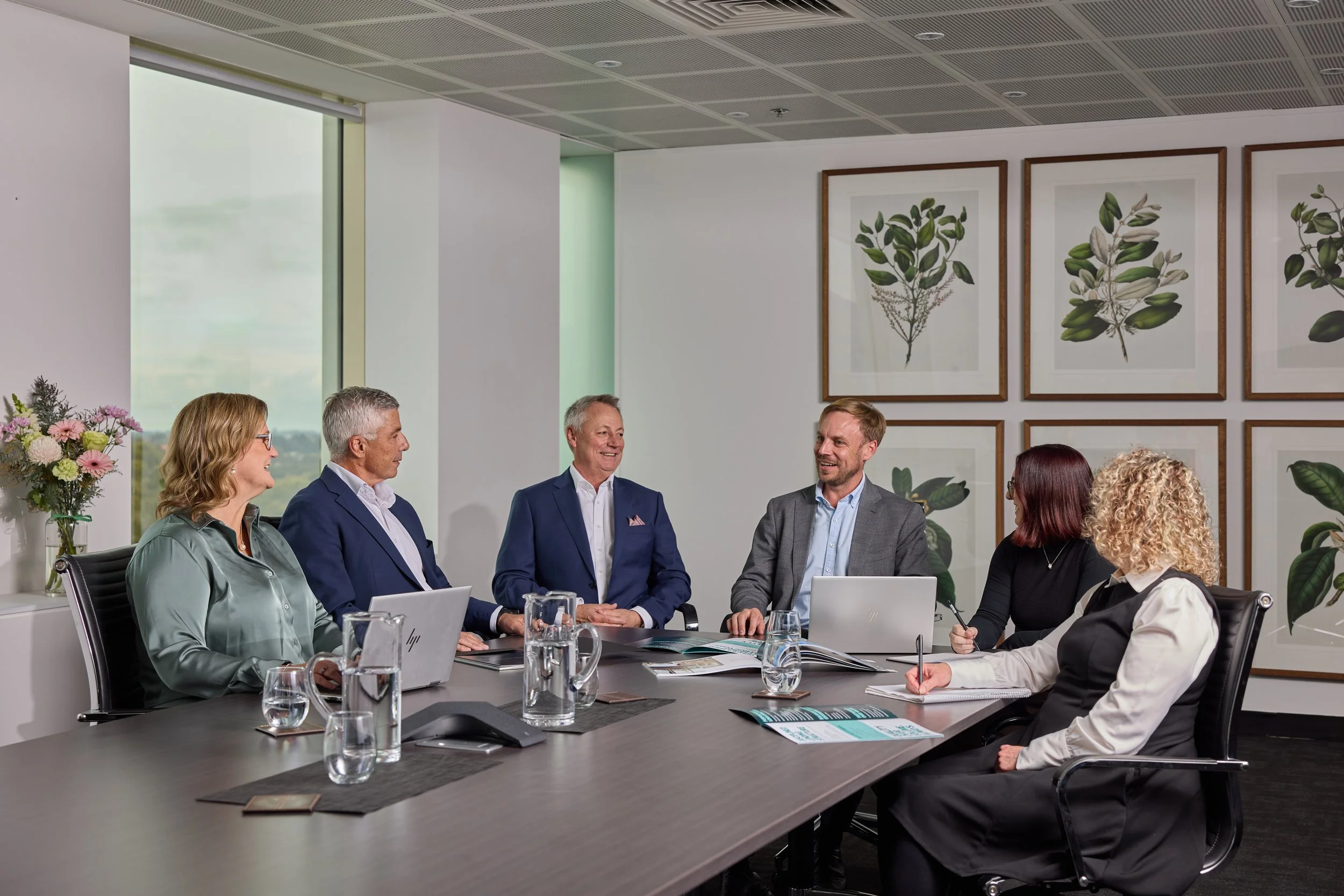 A group of professionals engaged in a meeting in a modern conference room with framed plant artwork and large windows.