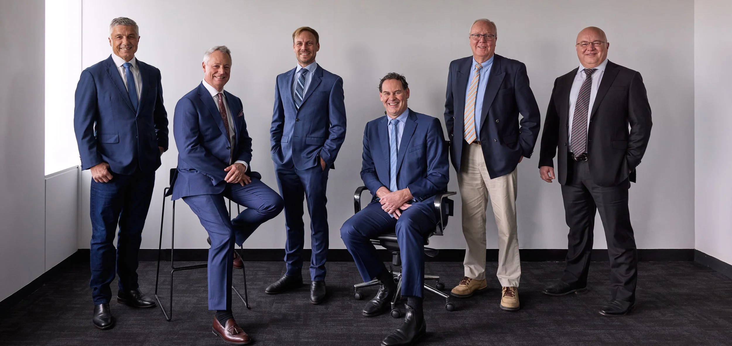 Group of seven men in business suits posing in an office room with white walls and black carpet, two are sitting and five are standing, all smiling.