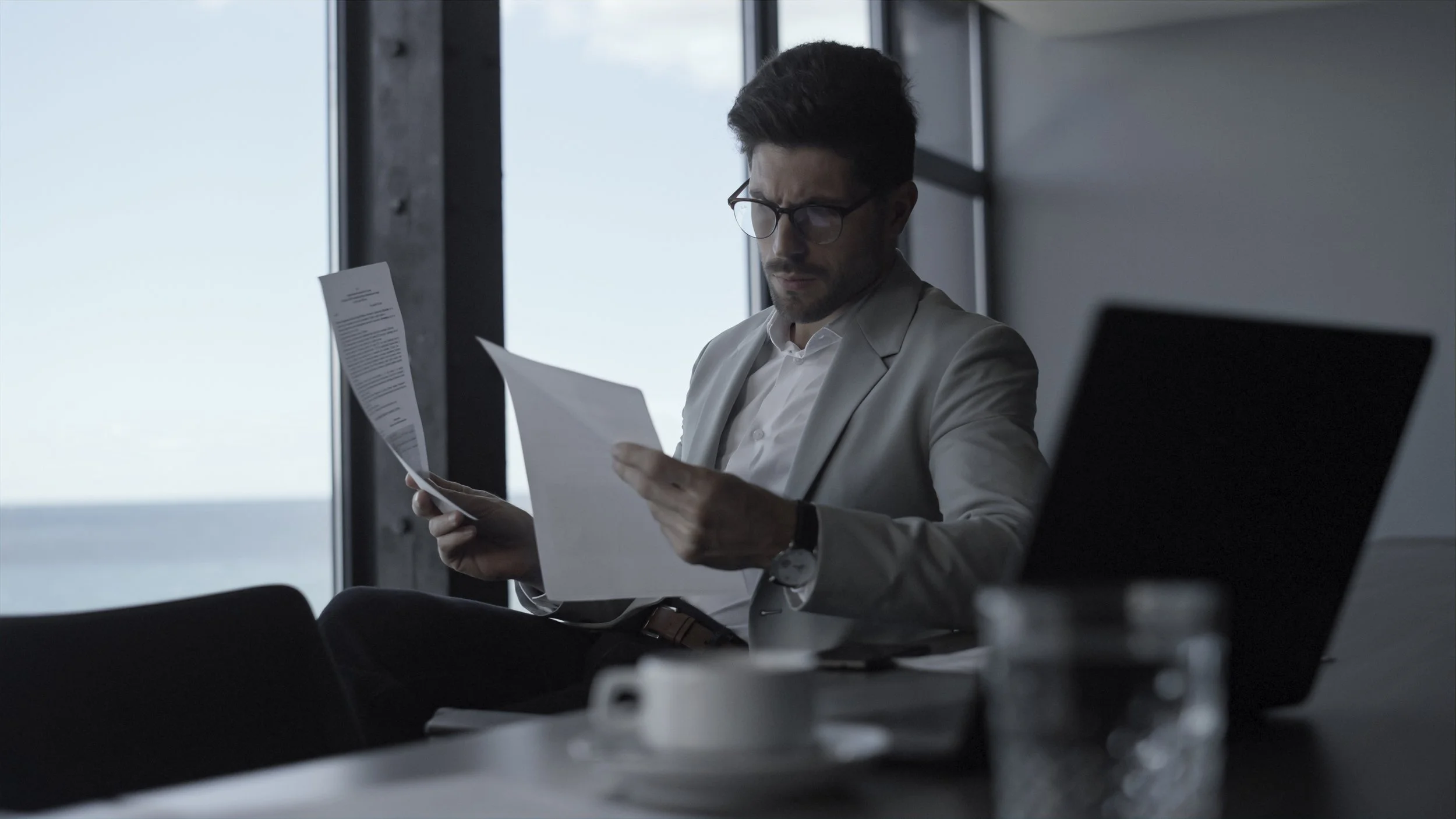 A man in a gray suit and glasses sitting in an office, reading a document with a serious expression.