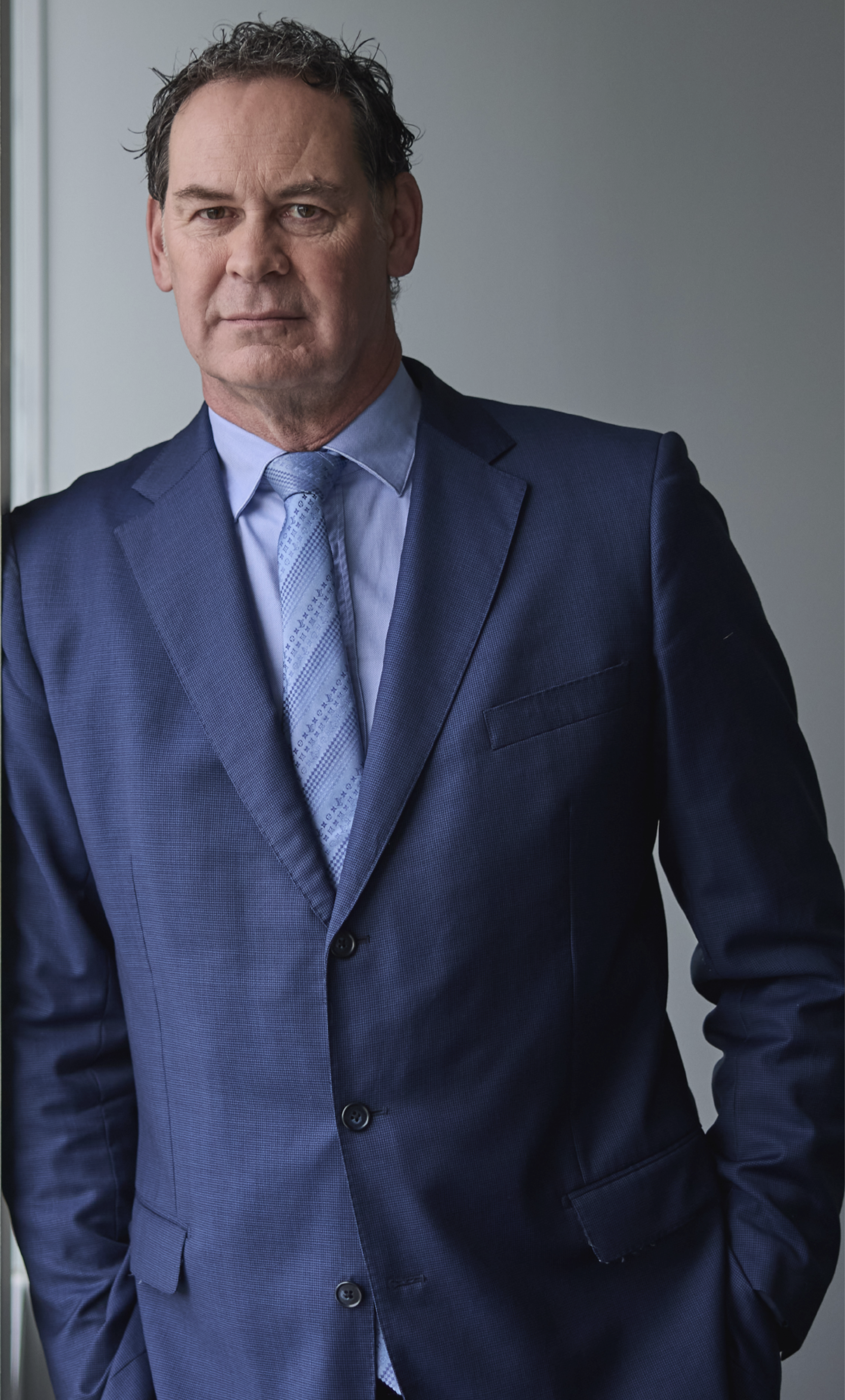 A man in a blue suit with a light blue shirt and patterned tie, standing indoors against a neutral background.