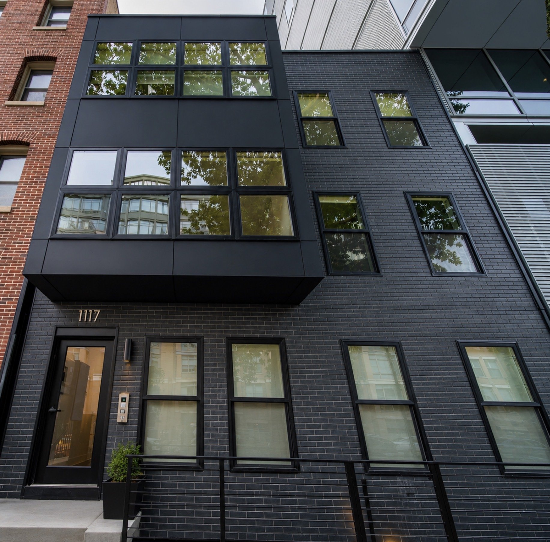 Modern multi-story building with black brick facade, large windows, a small green bush at the base, and a stairway leading to the entrance.