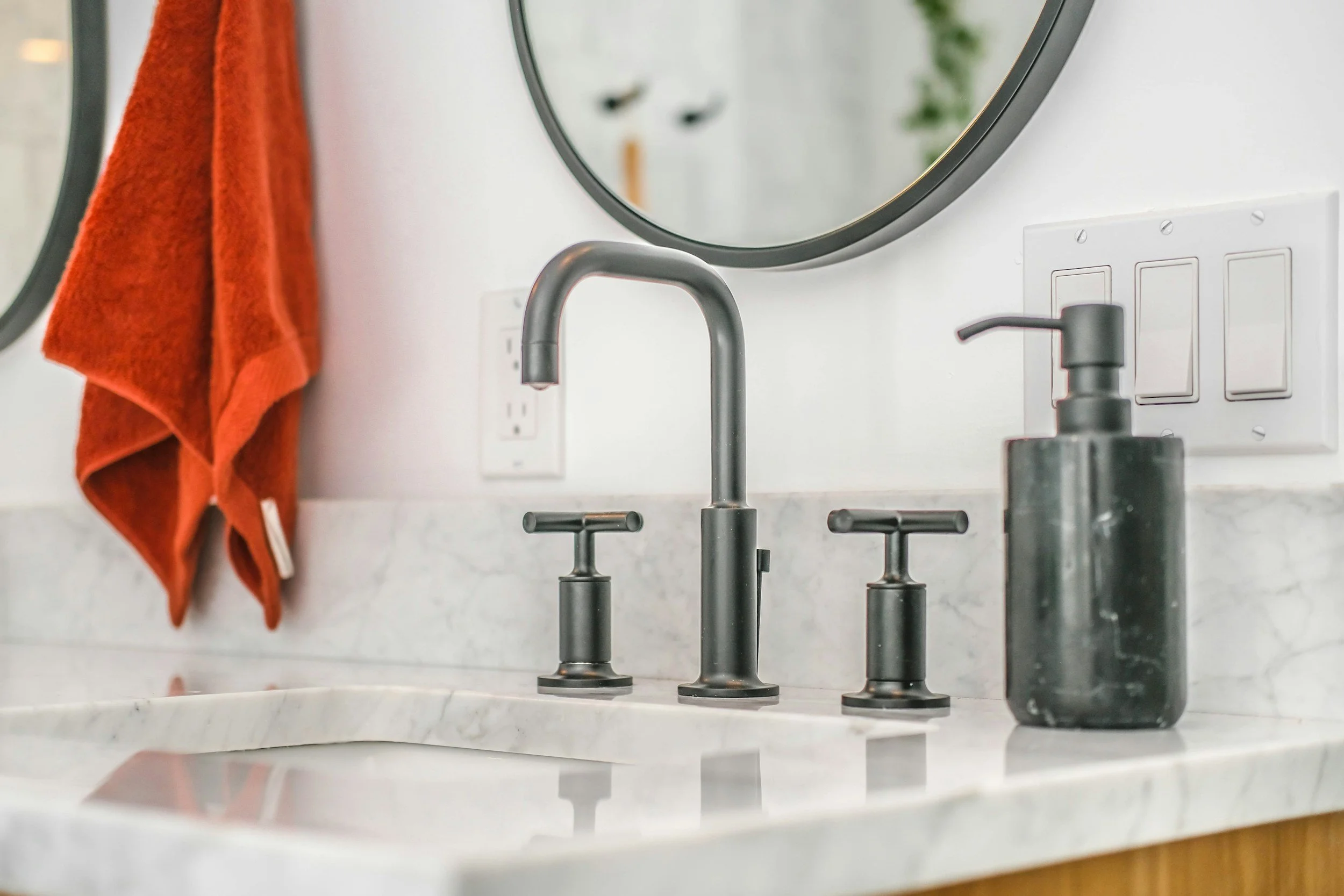 Close-up of a kitchen sink with two black faucets on a white marble countertop, with a mirrored wall above reflecting a plant and part of a face, and an orange towel hanging on a hook.