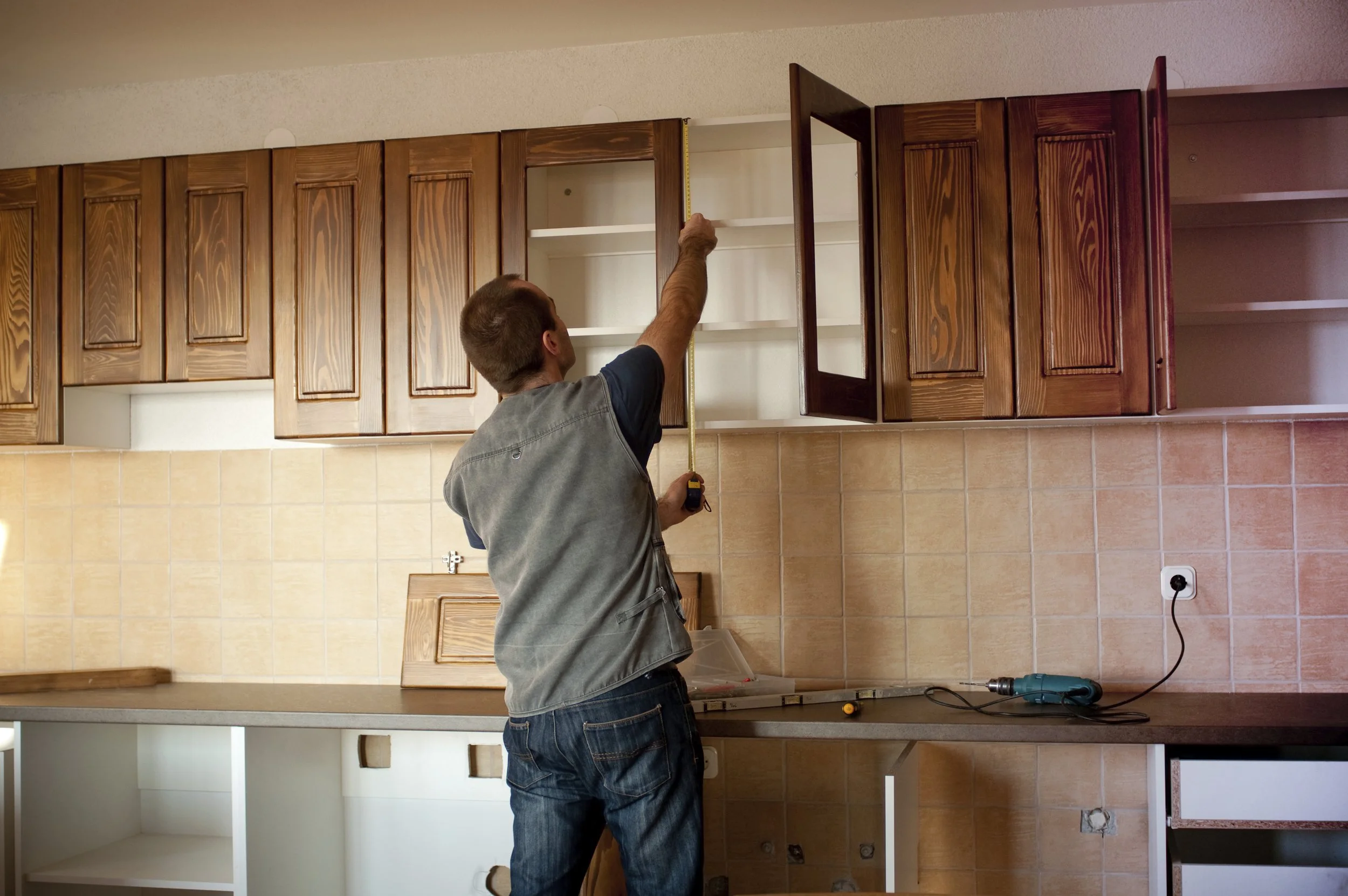 A man building or installing kitchen cabinets with a measuring tape, standing in a kitchen with beige tile backsplash and wooden upper cabinets.