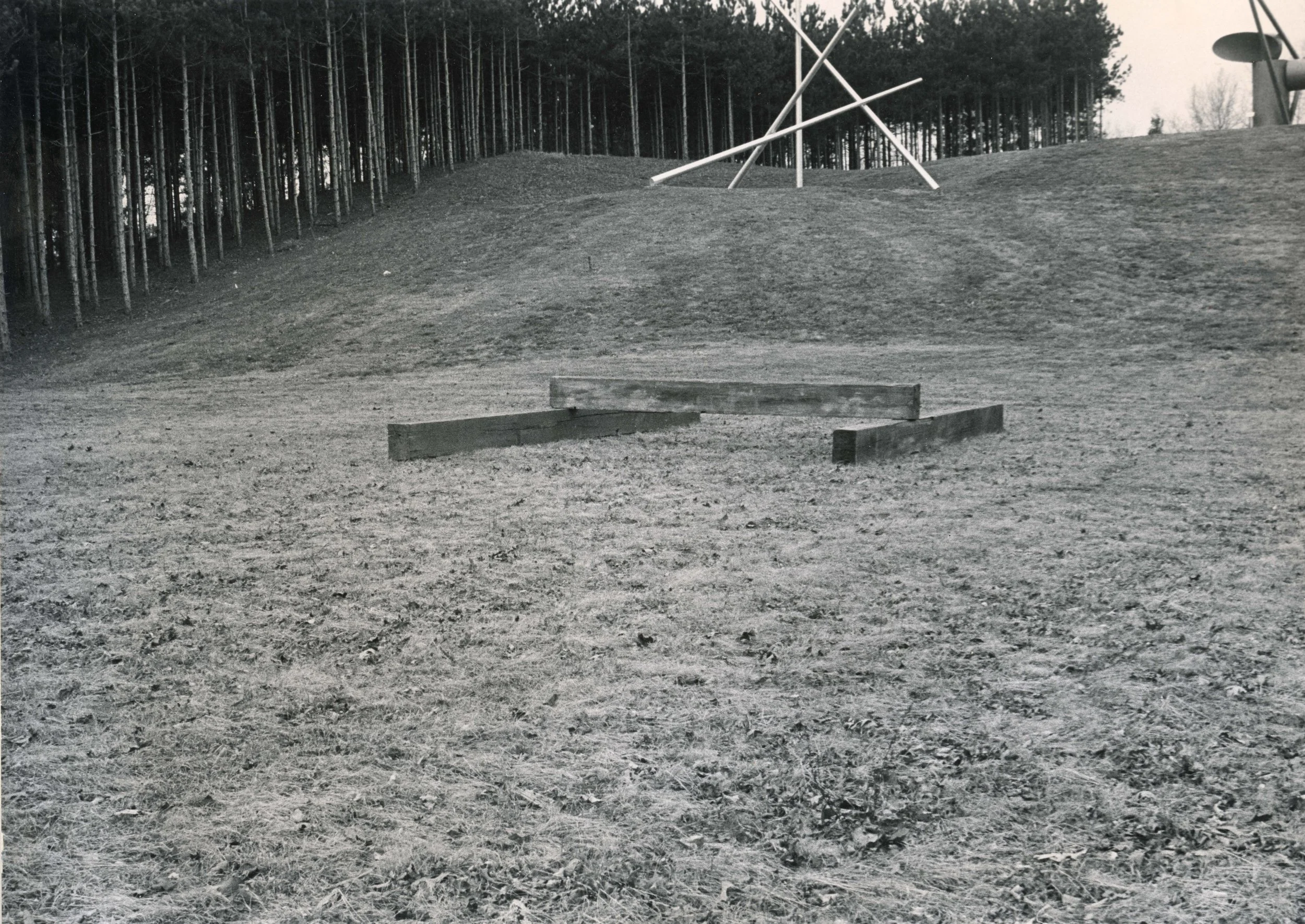 Black and white photograph of a grassy area with a wooden bench in the foreground, a hill with sculptures on top, and a forest of tall trees in the background.