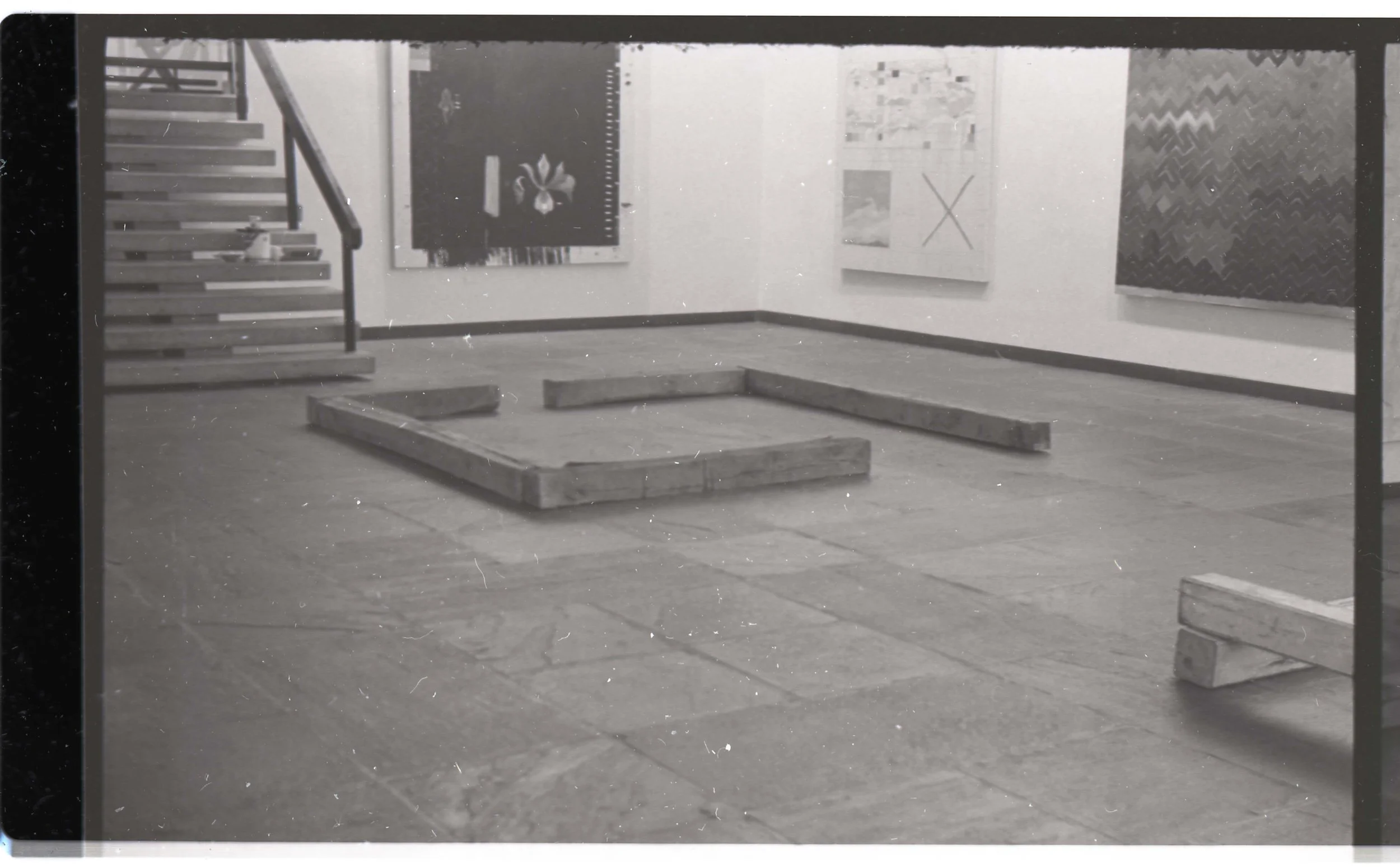 An art gallery with abstract paintings on the walls, wooden bench seating, and a staircase on the left side of the room.