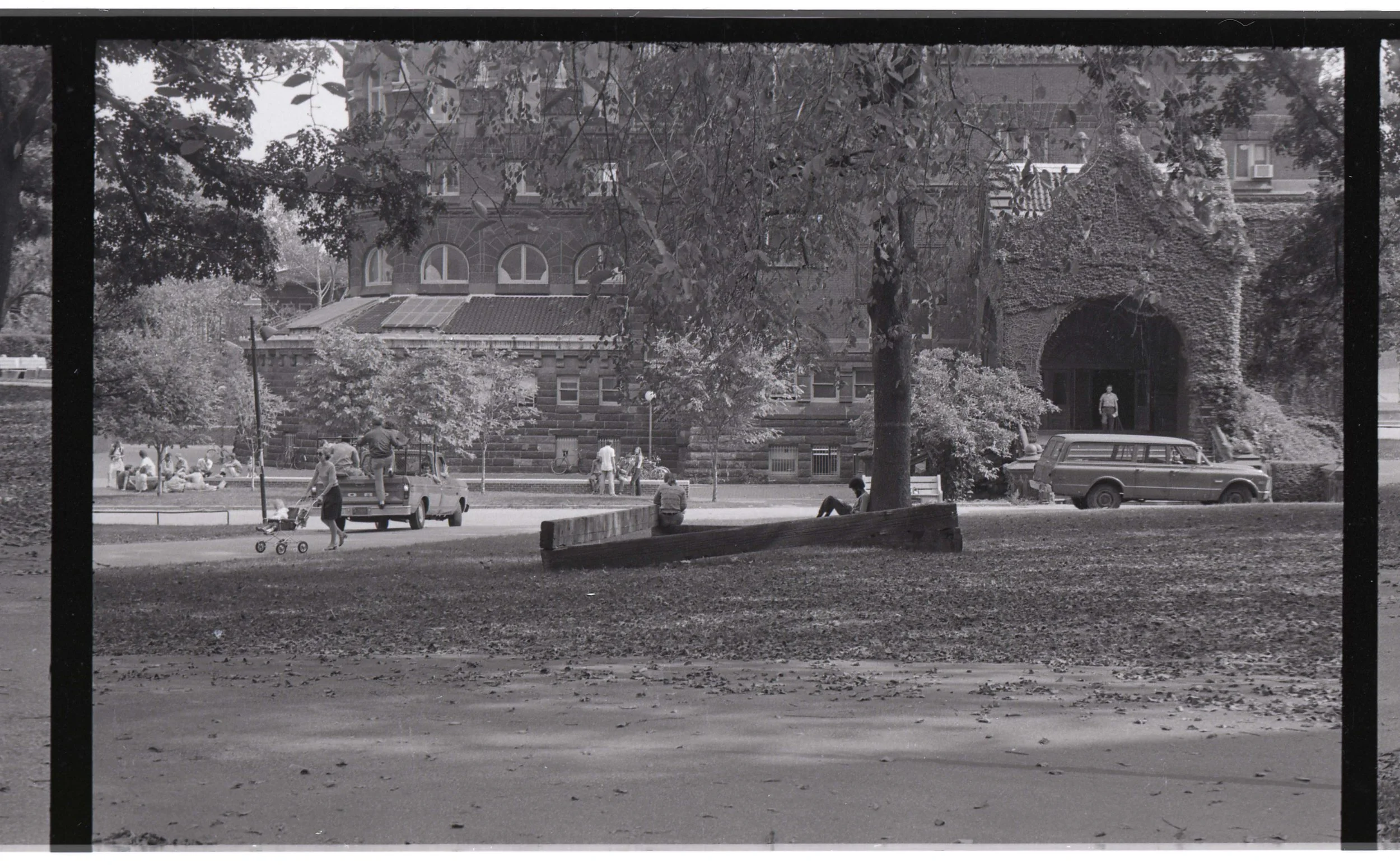 A black and white photograph taken from inside a frame or window of a park scene with people, trees, vintage cars, and a large stone building in the background.