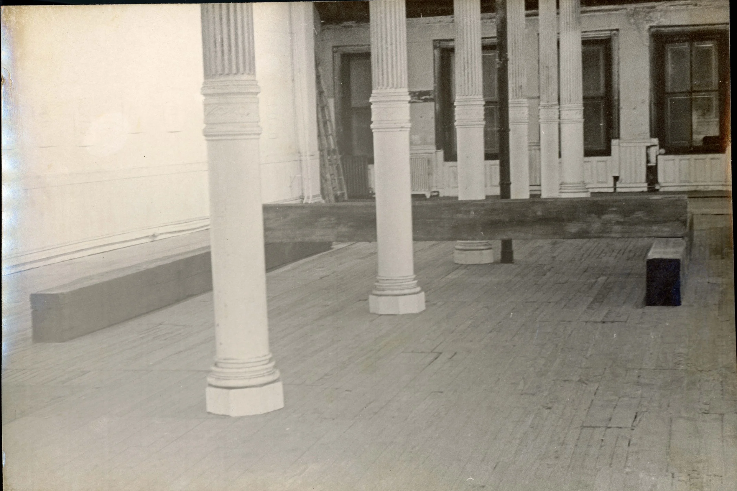 Black and white photo of a porch with wooden flooring, several white columns, and a home with multiple windows and a door in the background.