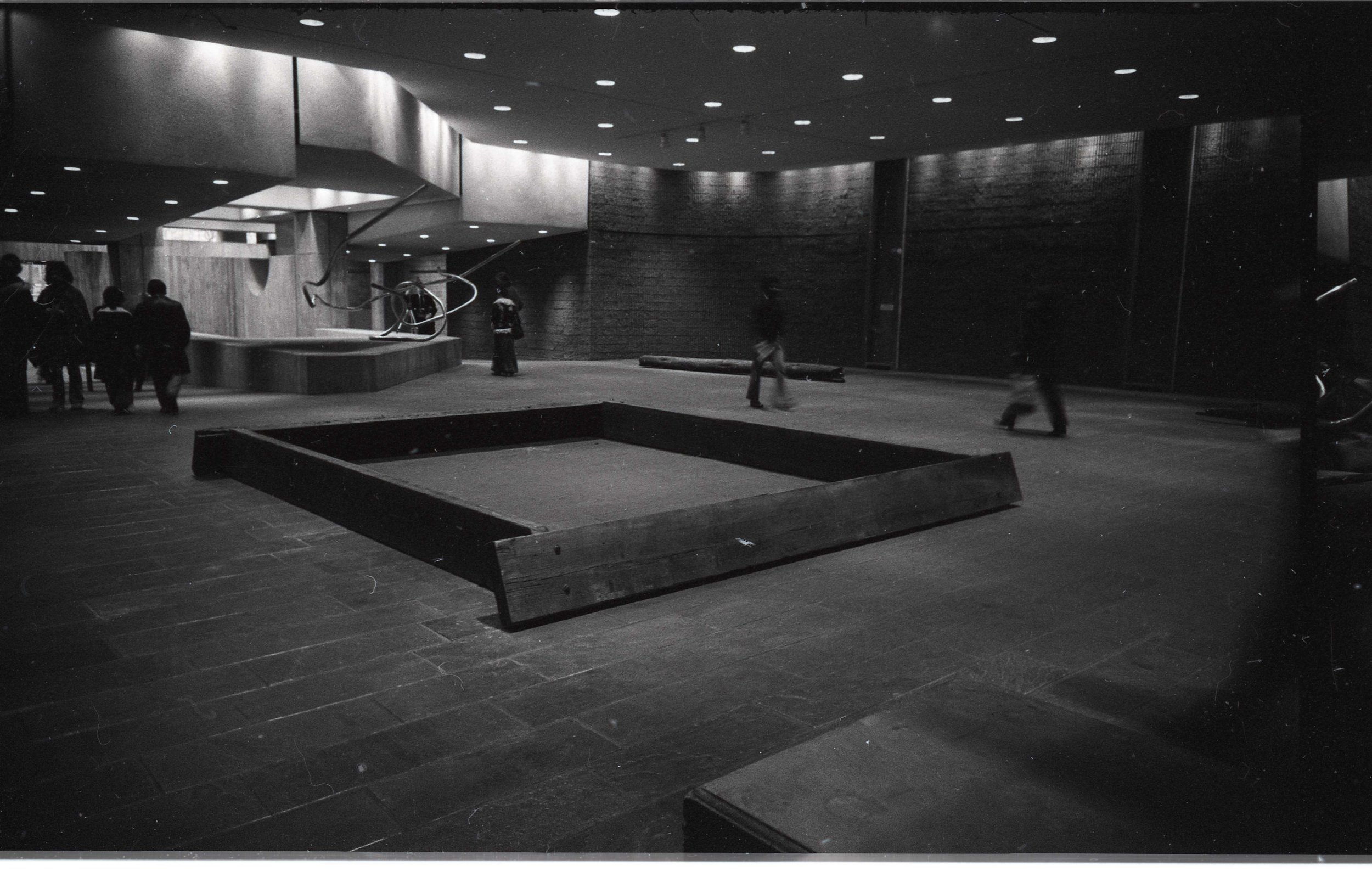 Black and white photo of people walking in a modern, minimalist indoor space with a curved wall, recessed lighting, and an art installation on a stand.