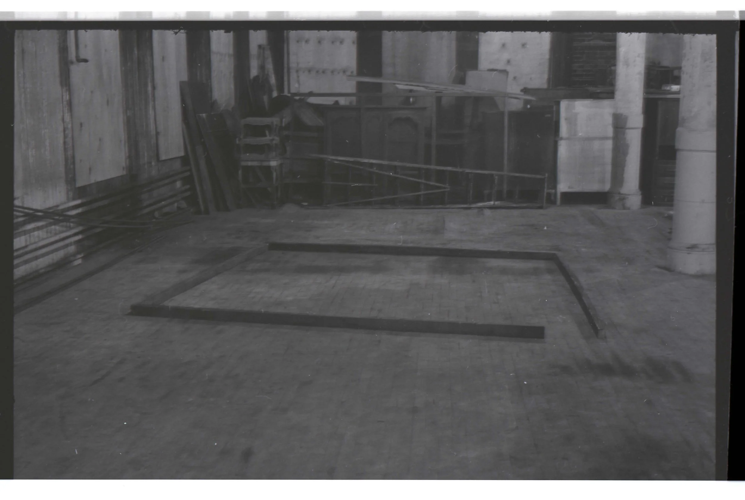 Black and white photo of an empty room with wooden walls and floor. A metal roof is visible overhead. There is a shelving unit or scaffolding and some chairs stacked in the corner. Two large concrete columns support the roof.