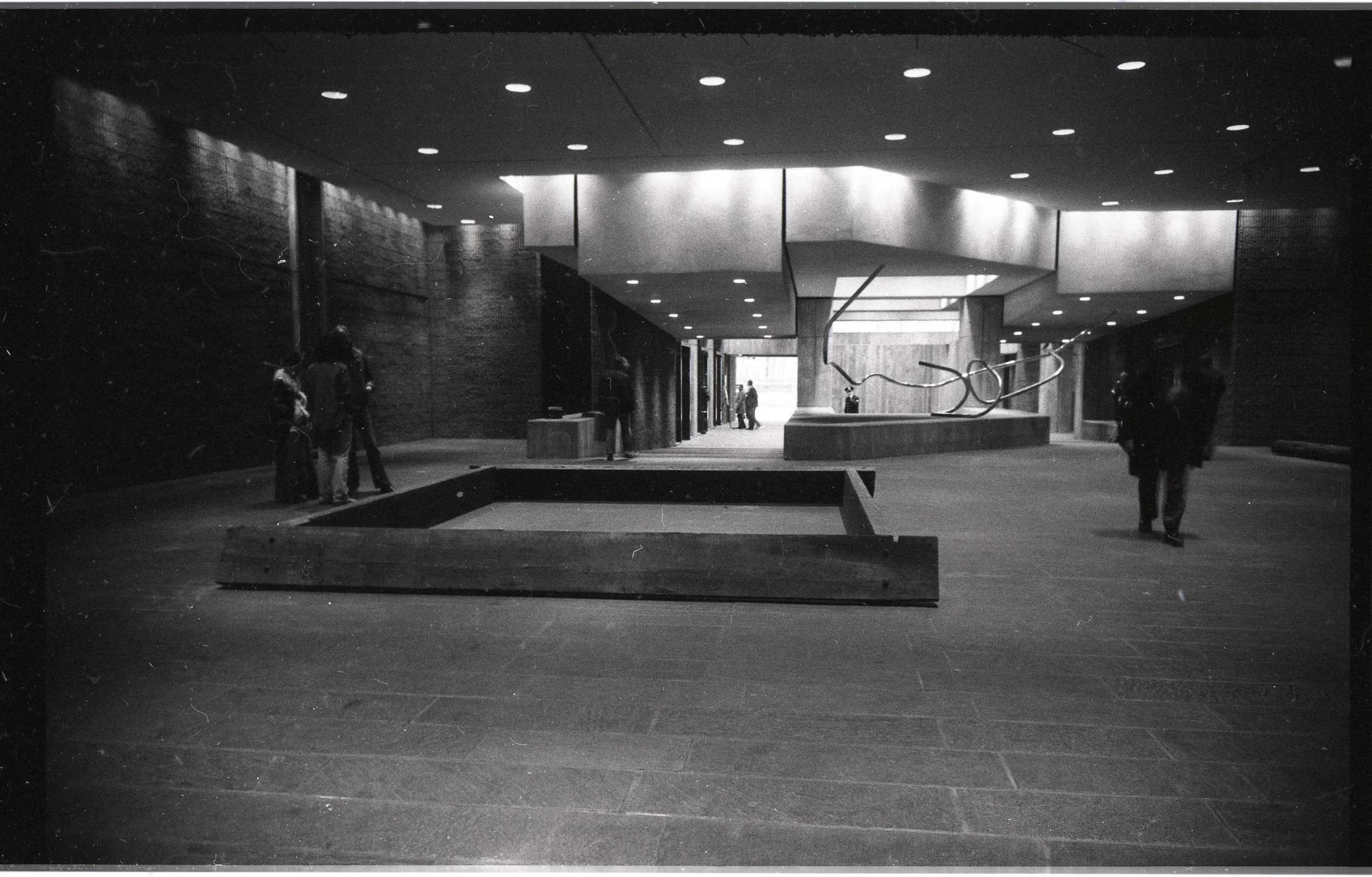 Black and white photo of the interior of a modern building with a high ceiling and multiple ceiling lights. A few people are standing or walking near the walls and an abstract metal sculpture is visible in the foreground. The architecture features co