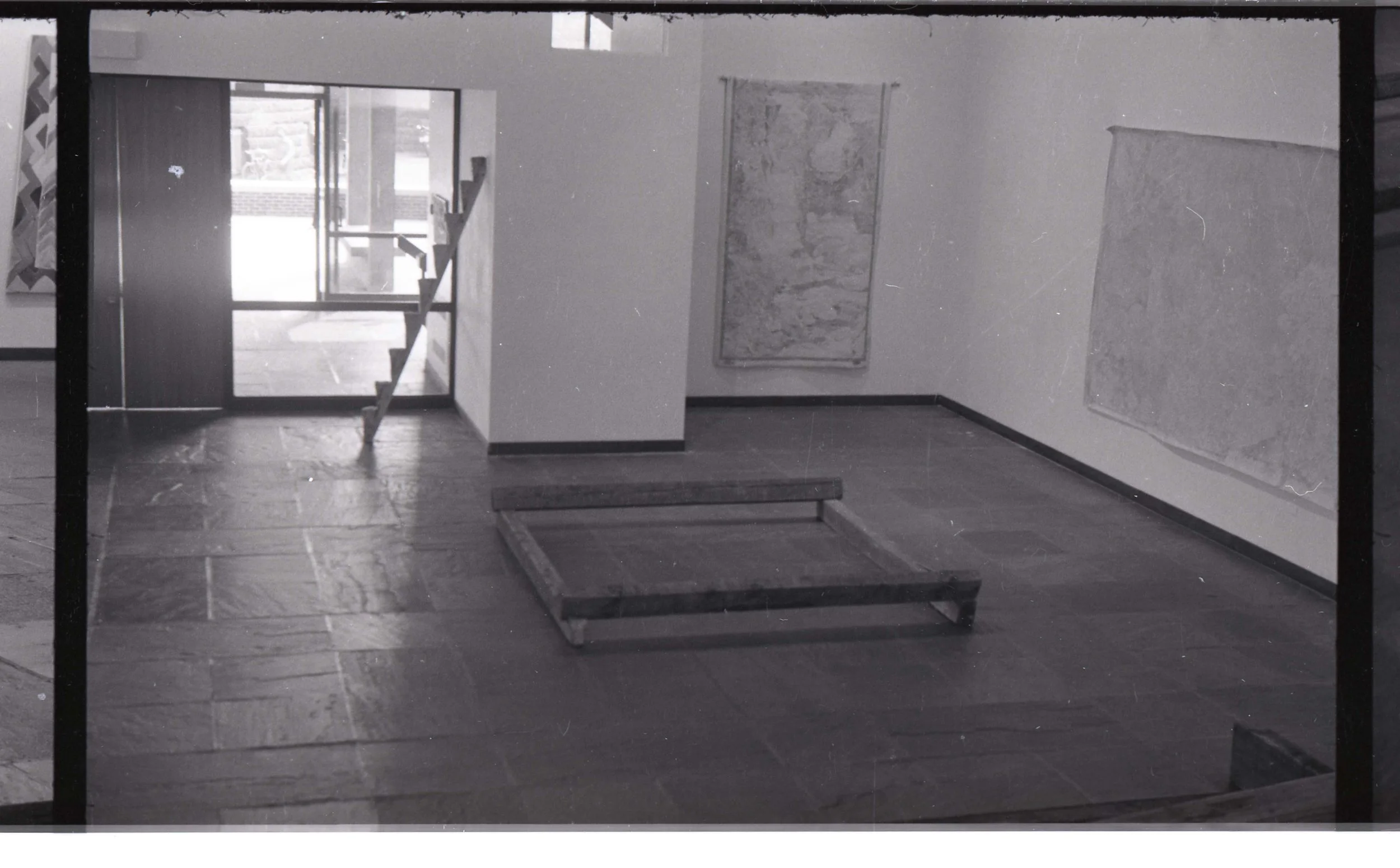 Empty room with tiled floor, two picture frames on the wall, and a partial staircase with a railing leading to an upper level. Front door with glass panels in the background.