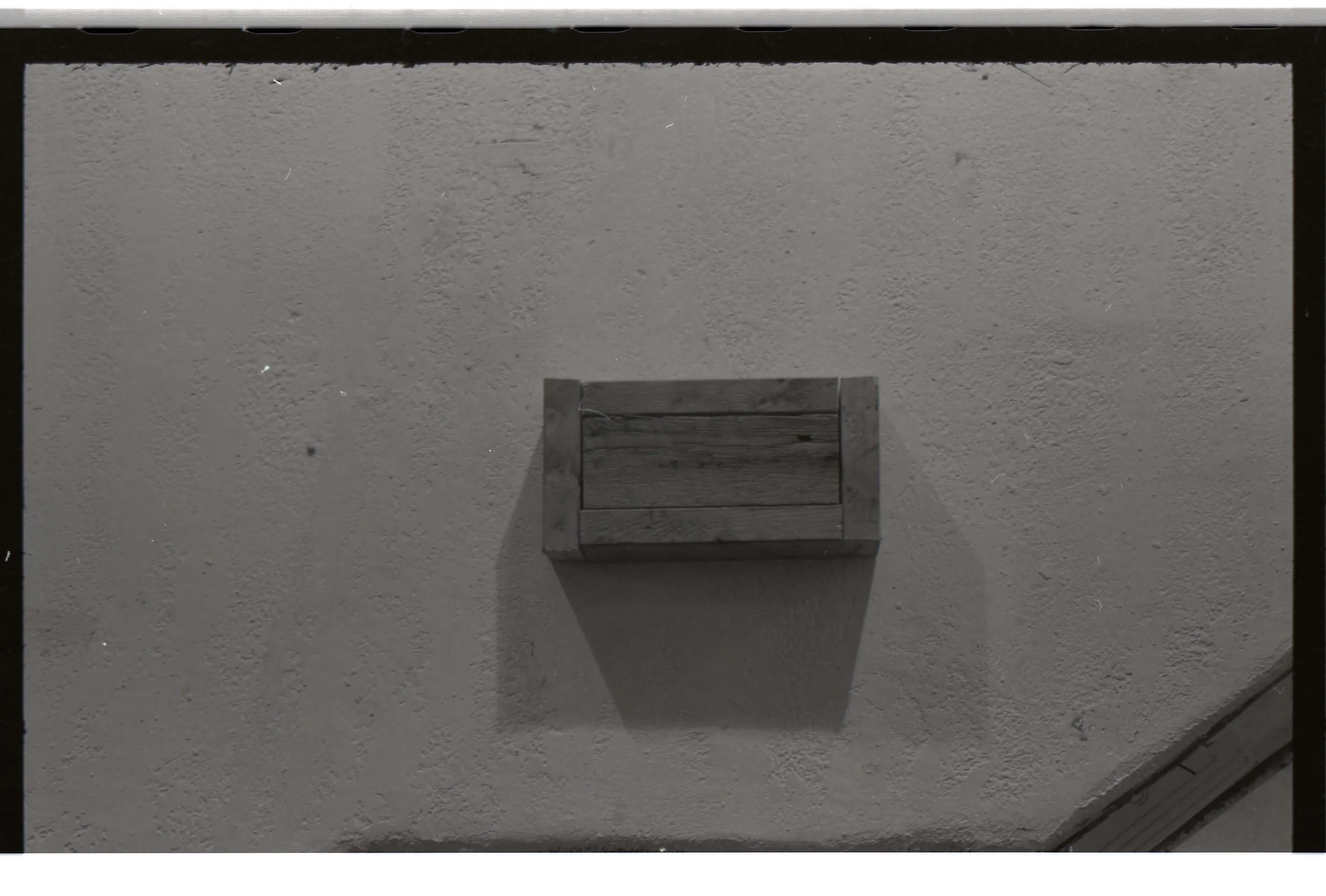An outdoor scene showing a textured gray wall with a small wooden box mounted on it and a shadow cast below the box.