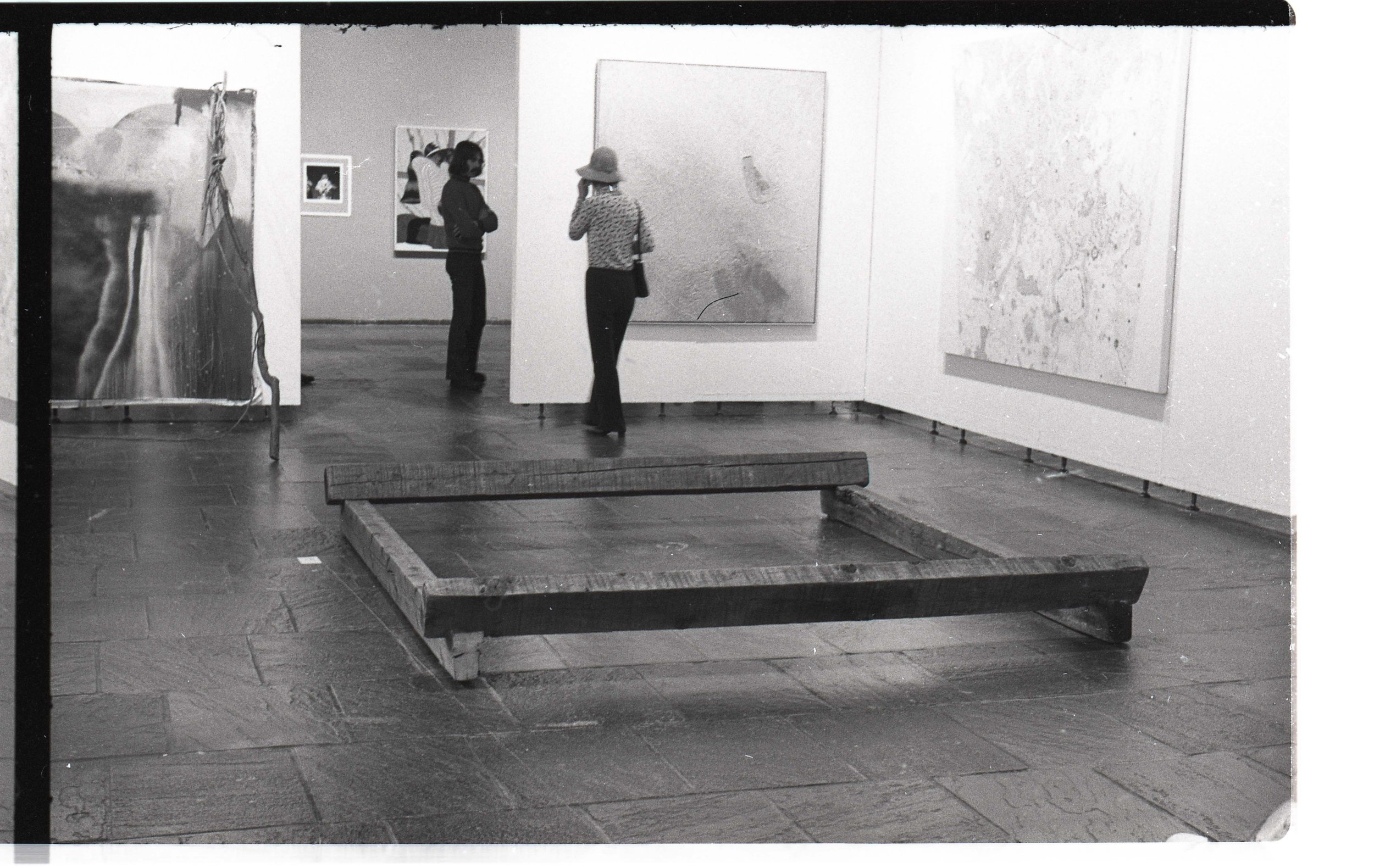 Two women are looking at artwork in an art gallery. One woman is wearing a coat with her arms crossed, and the other is wearing a hat and patterned shirt. Artworks are hanging on white walls behind them.