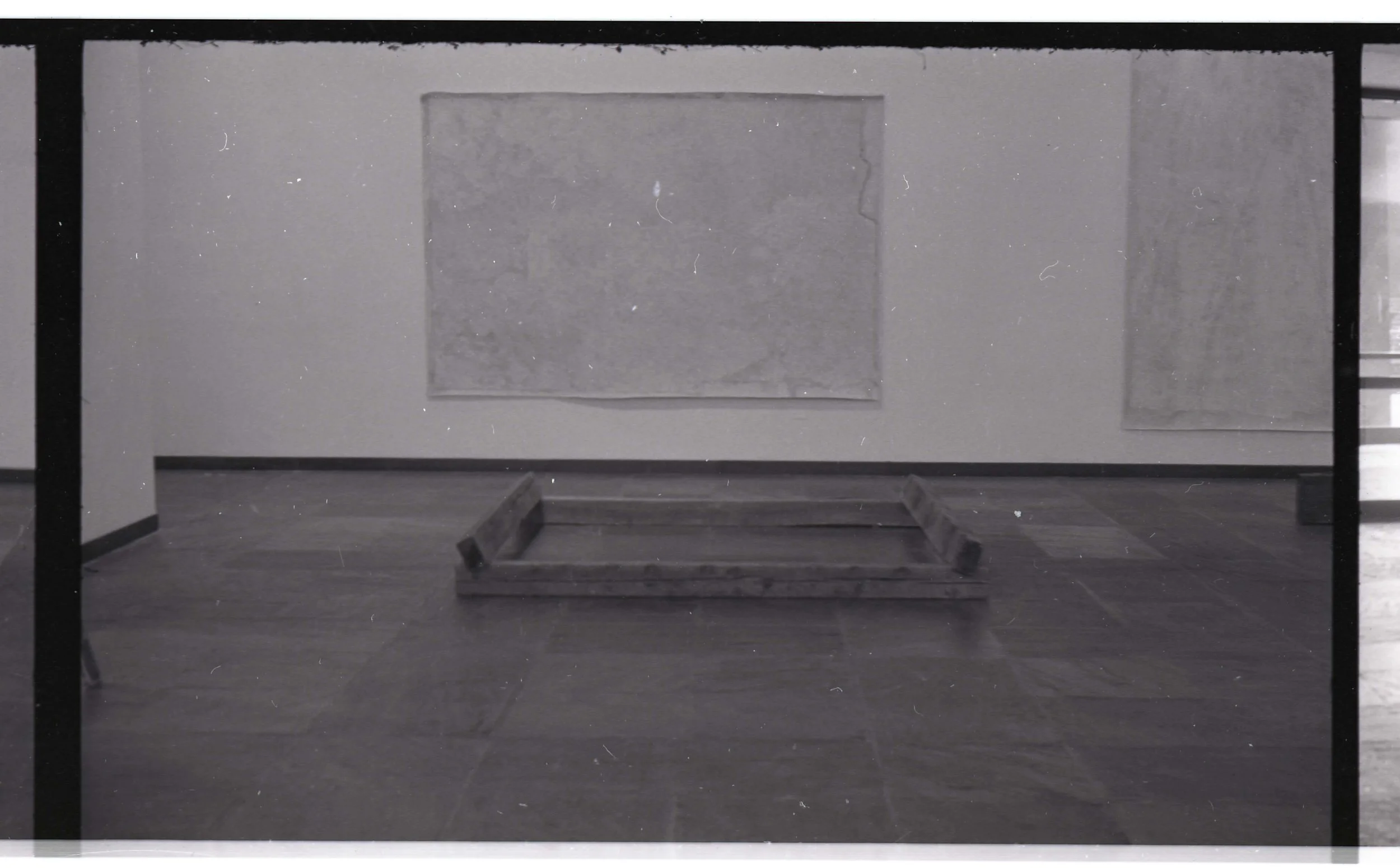 An outdoor scene with a rectangular wooden frame on a concrete surface, a wall with a square opening, and a partially snow-covered ground.