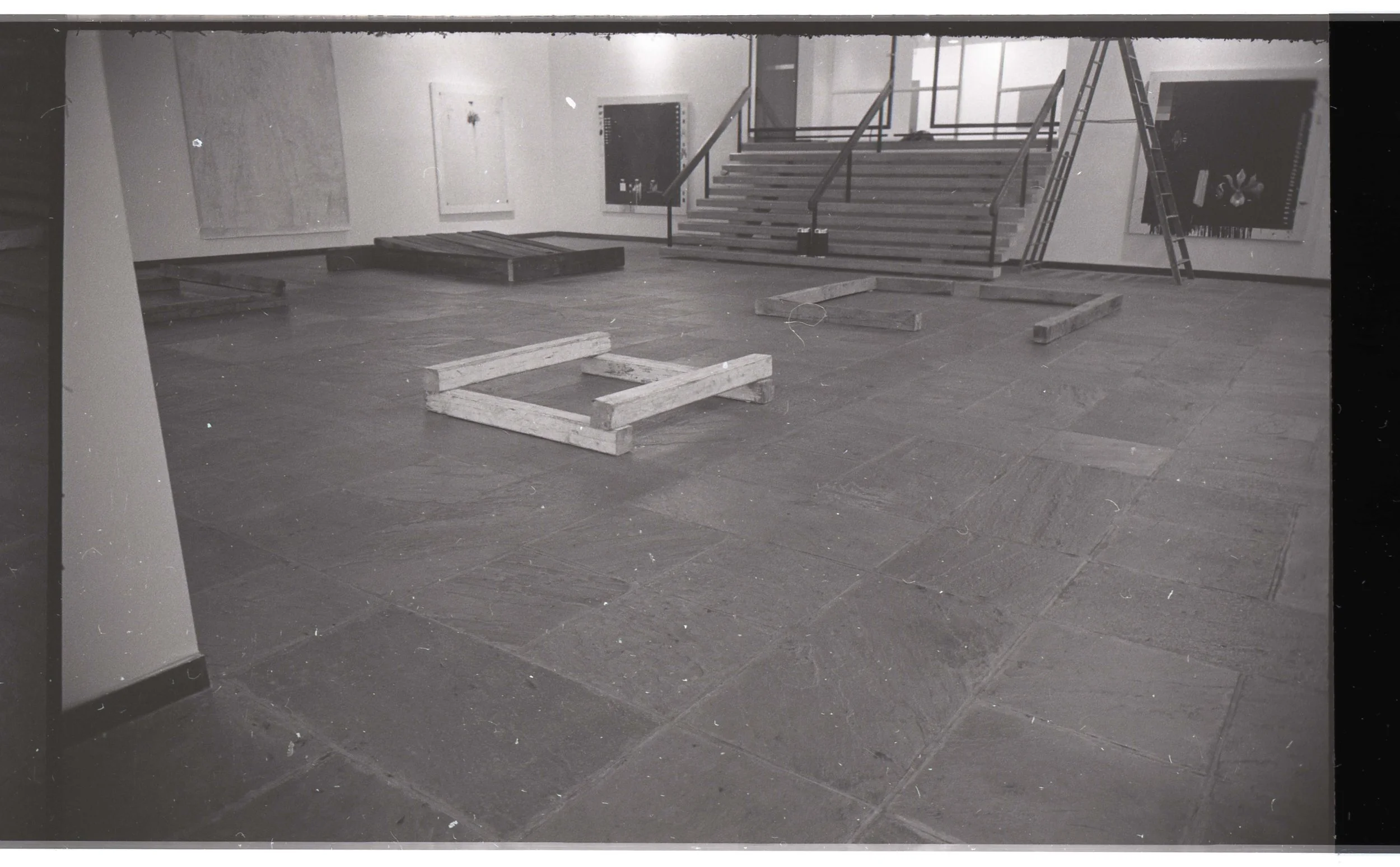An art gallery or museum room under construction with wooden frames on the floor, a staircase leading to an upper level, and artworks on the walls.