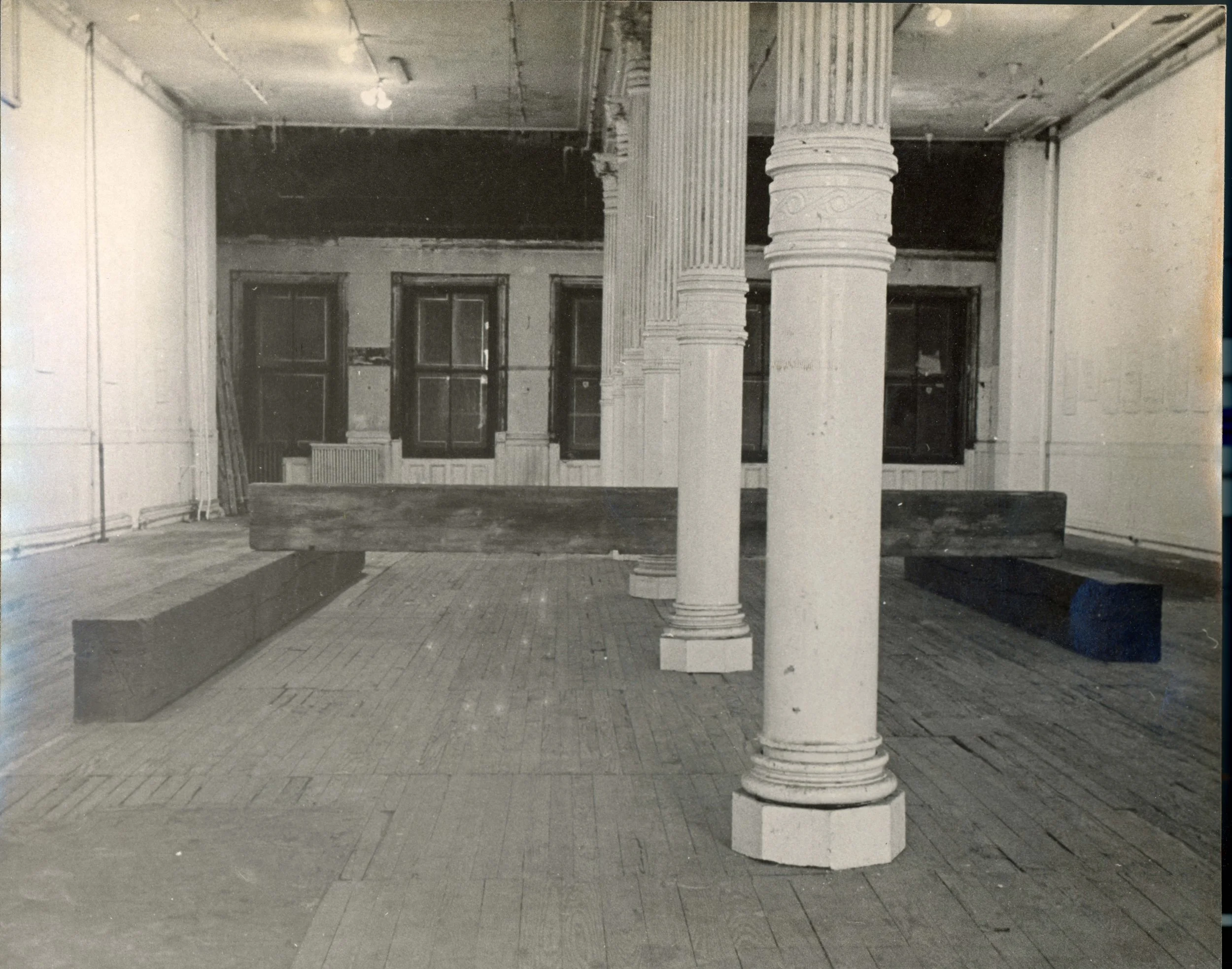 Black and white photo of an empty room with a tile floor, large windows, and a ceiling with visible light fixtures. There are two prominent decorative columns in the center of the room. Some furniture is visible, including a bench or table in the for