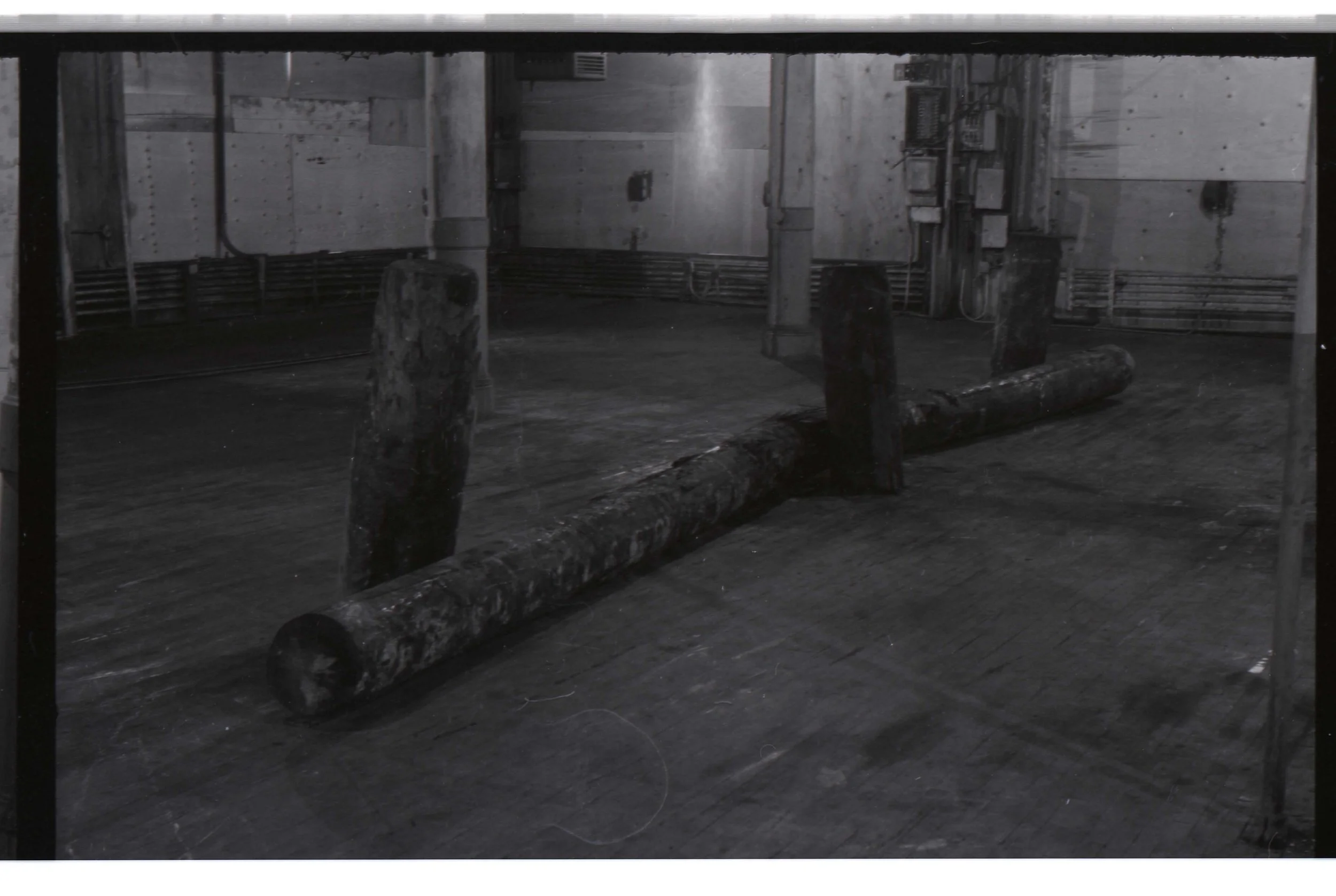 Black and white photograph of an industrial space with wooden beams, pipes, and electrical boxes. Two large logs are placed on the floor, one upright and the other horizontal.
