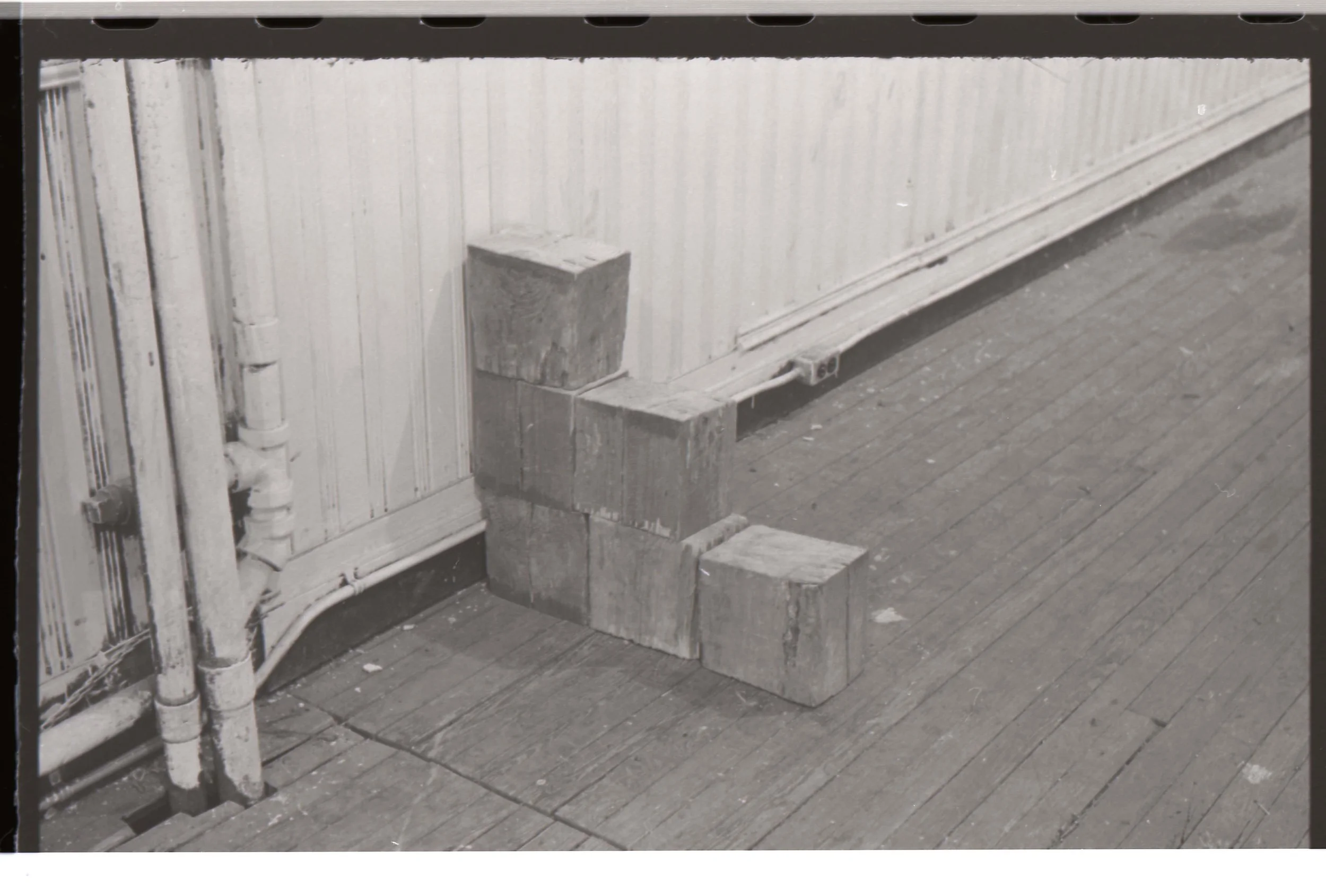 Black and white photograph of wooden stairs with three blocks of wood stacked at the bottom, next to a wall with pipes and electrical wires.