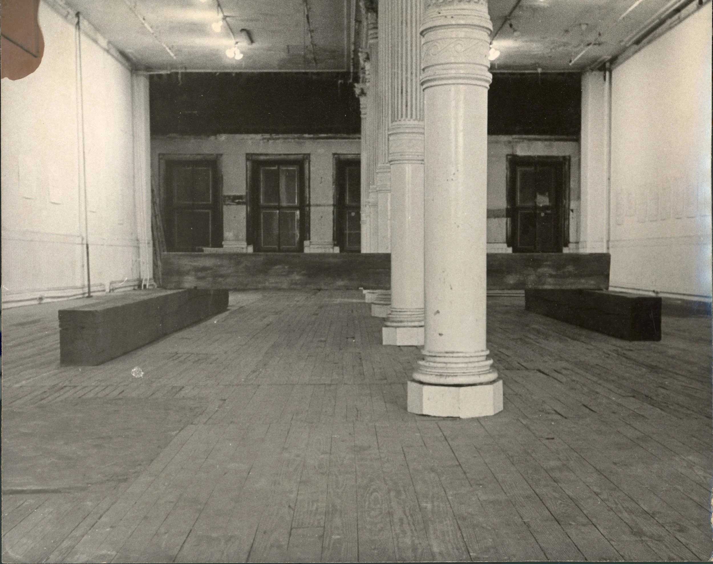 An empty room with brick flooring, white walls, and a large white column in the center. There are windows at the back and benches on either side. The ceiling has exposed pipes and a few hanging lights.