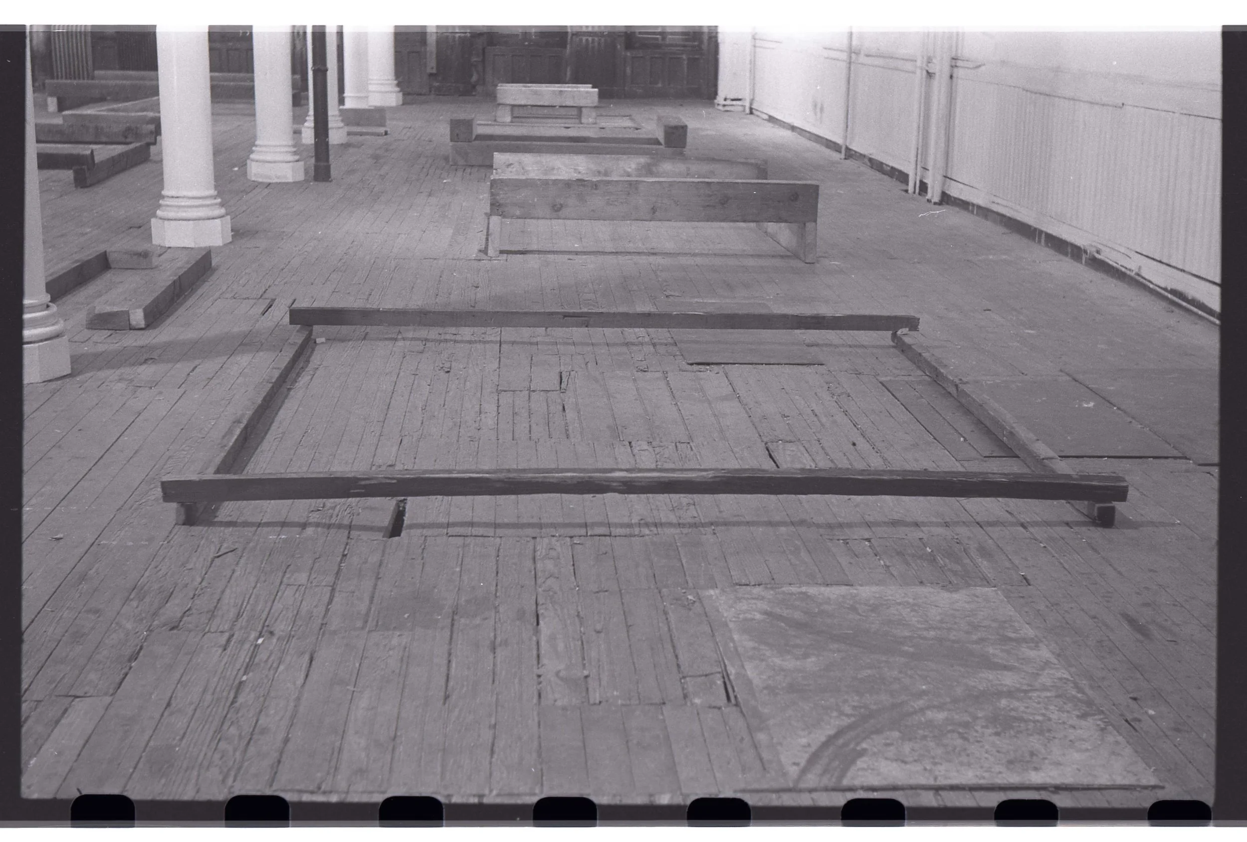 Black and white photo of unfinished wooden framework on a brick floor, with some vertical white columns and a wall with pipes in the background.