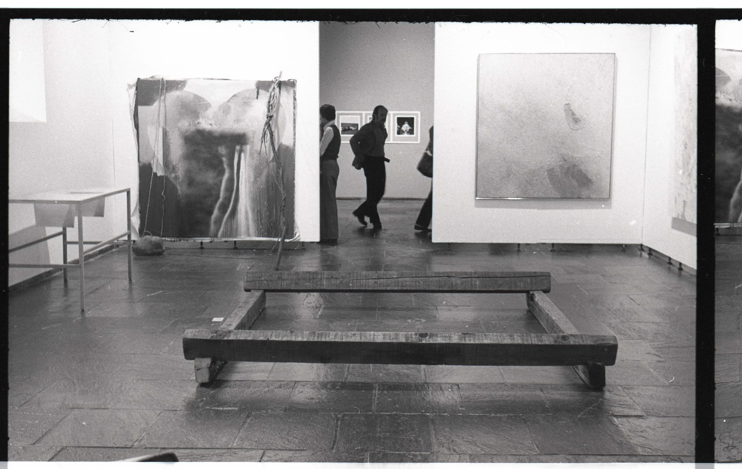A black and white photograph of an art gallery with several paintings on the walls. In the foreground, there are wooden benches and a small table. Several people are inside the gallery, looking at the artwork.