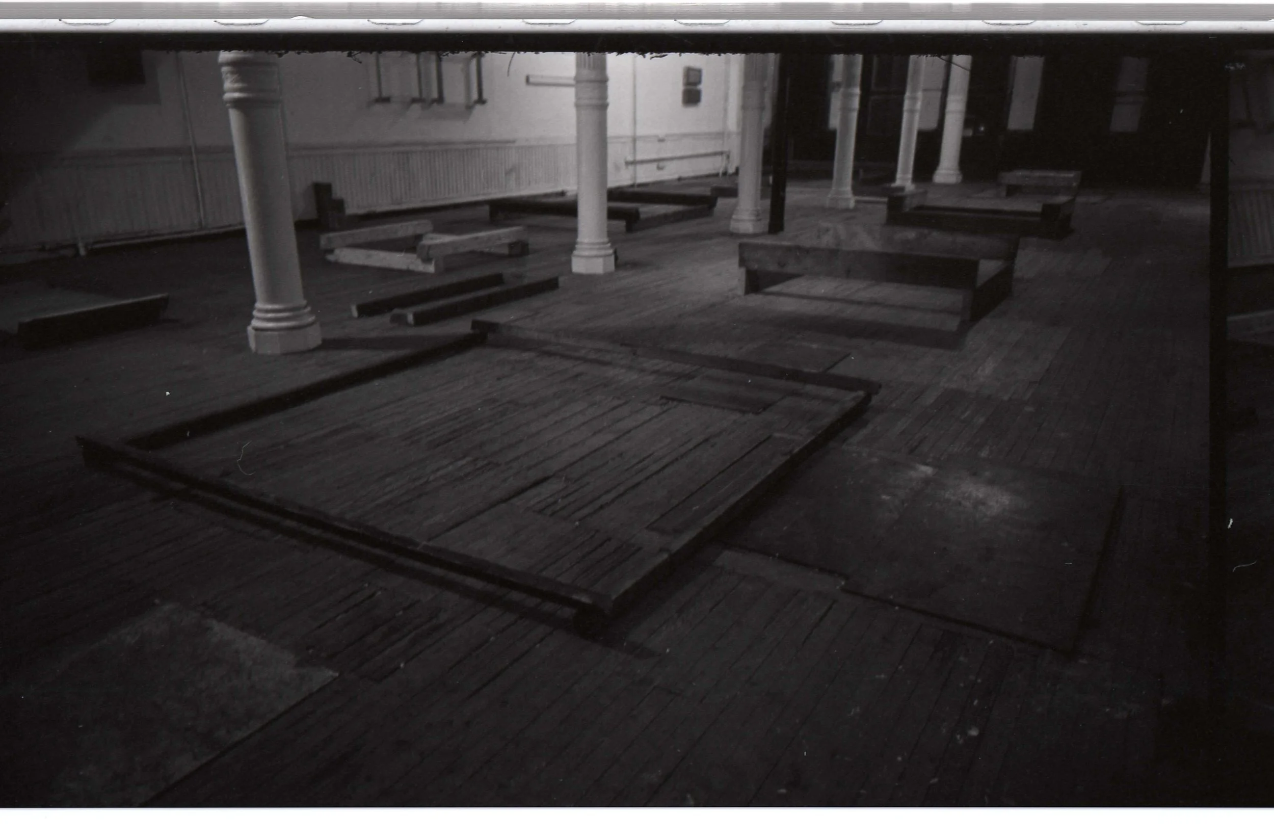 A dark, empty room with wooden floors, white columns, and scattered wooden benches, some toppled over.