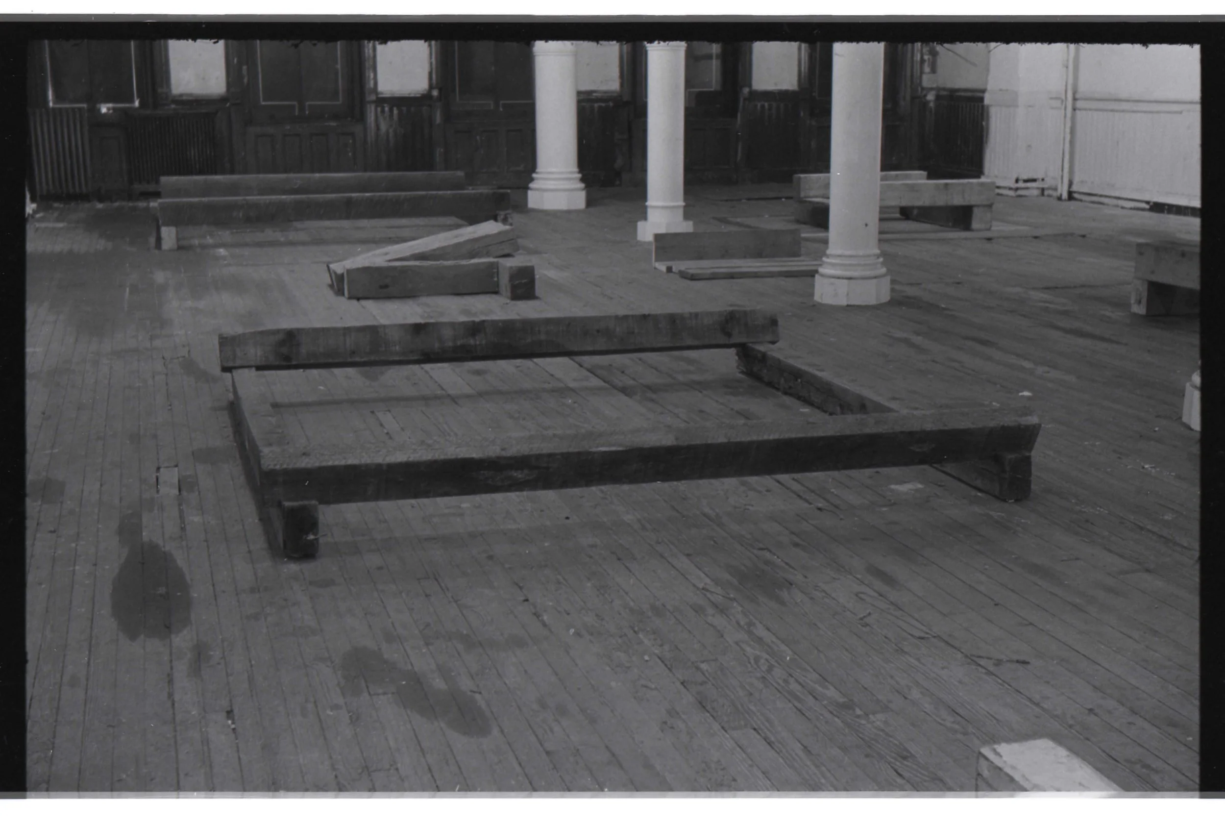 Empty wooden floor with scattered wooden planks and benches in an old-fashioned room with dark wooden paneling and white pillars.