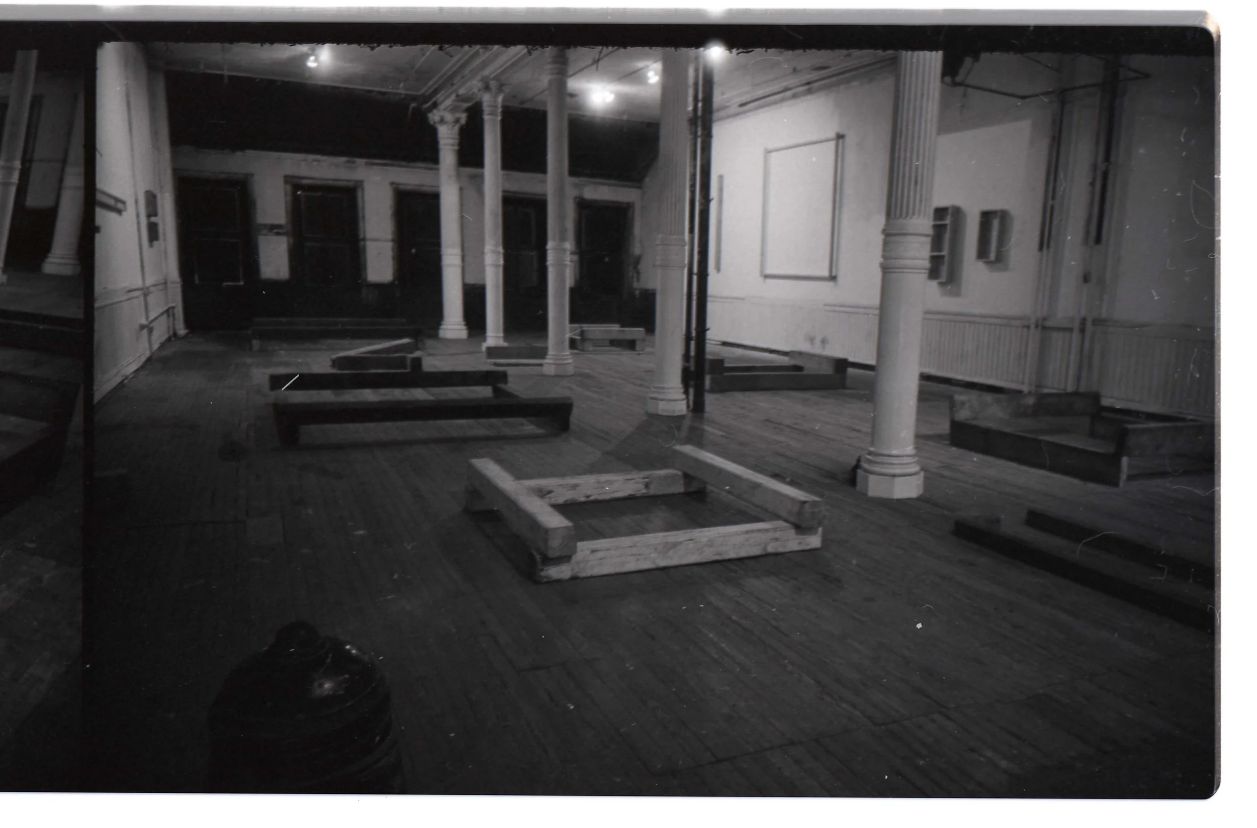 Empty room with wooden floors, partially assembled furniture frames, and white columns supporting the ceiling, with some walls and windows visible.