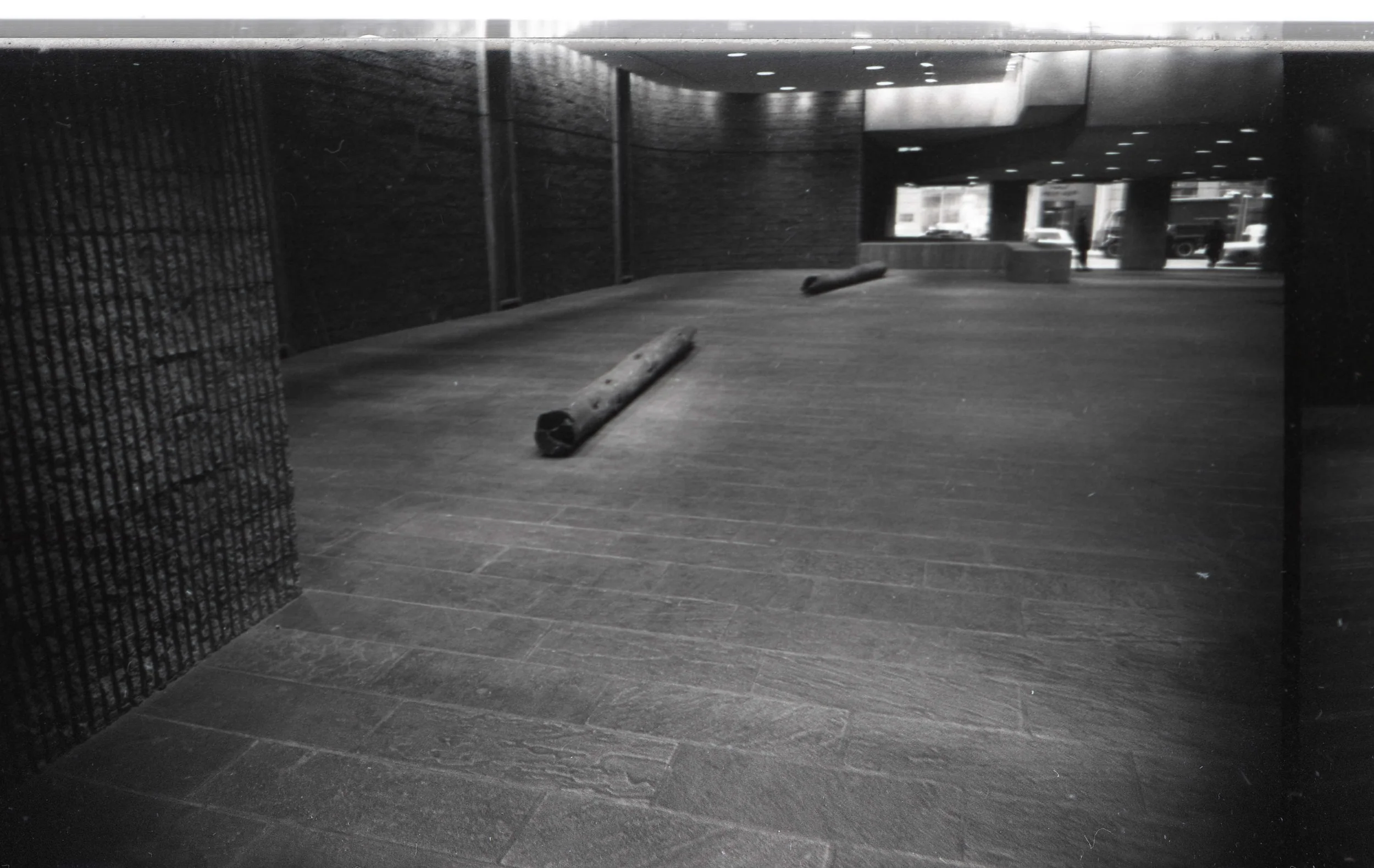 Black and white photo of an urban or commercial area with a tiled floor, a curved wall, large windows, and some large logs on the ground. In the background, a few parked cars and pedestrians are visible outside.
