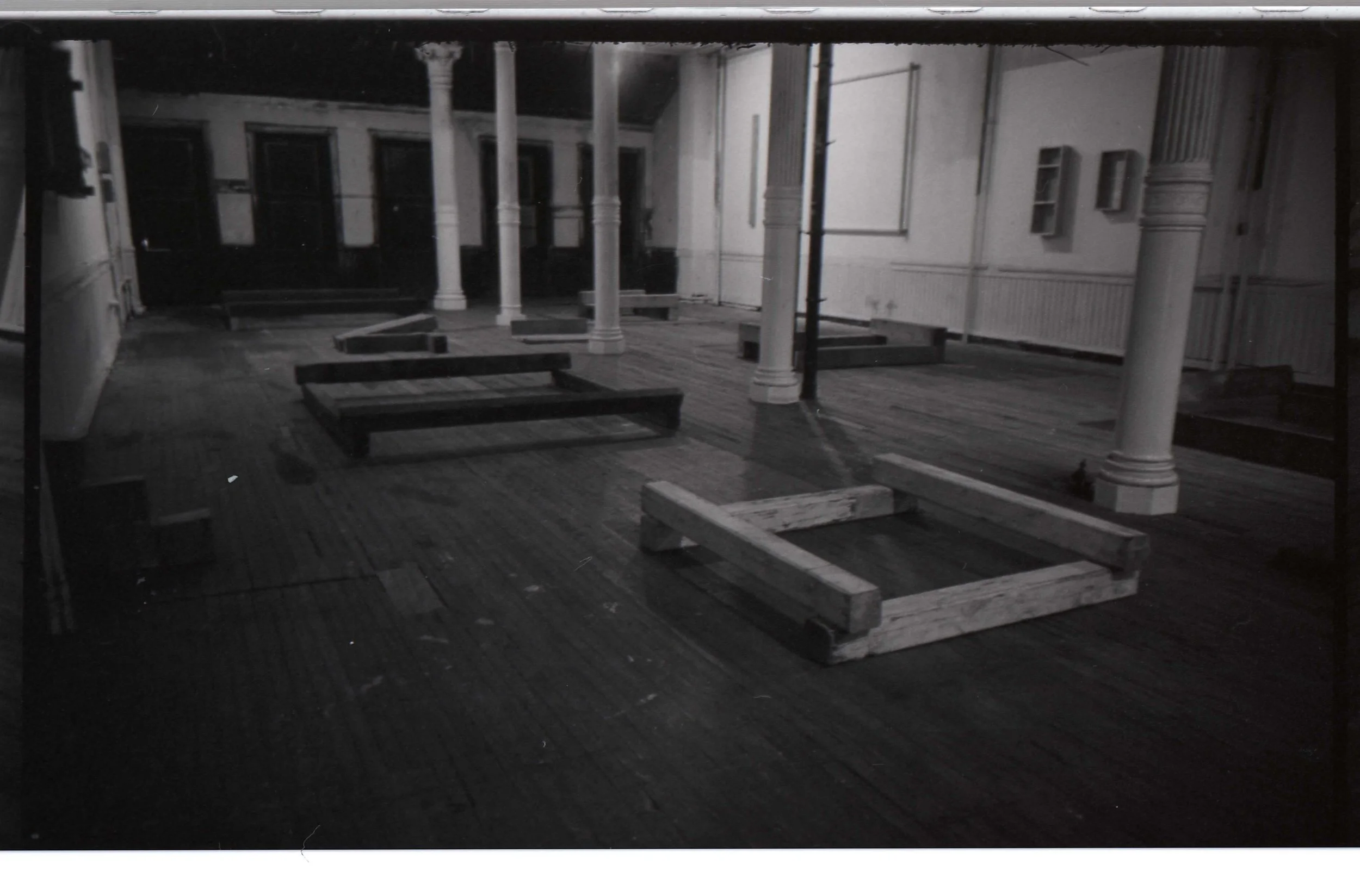 Black and white photo of an empty room with columns, window frames, and wooden flooring, with some wooden frames and structures on the floor.