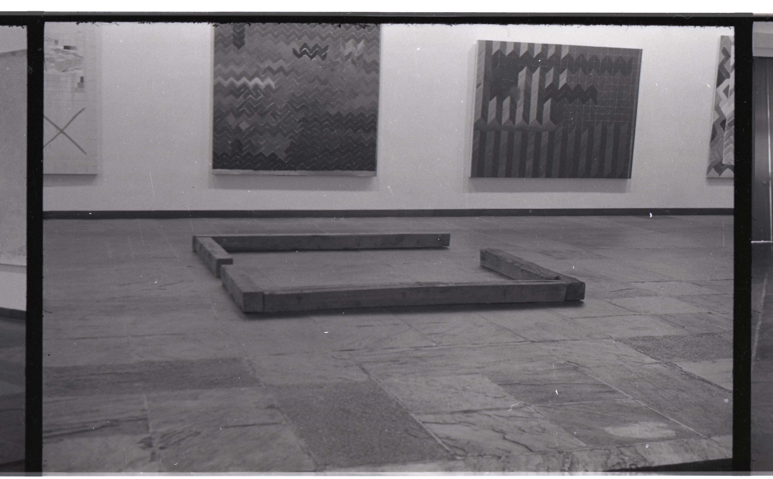 An art gallery with four framed abstract paintings on a white wall, a black metal railing in the foreground, and a wooden platform on the floor.