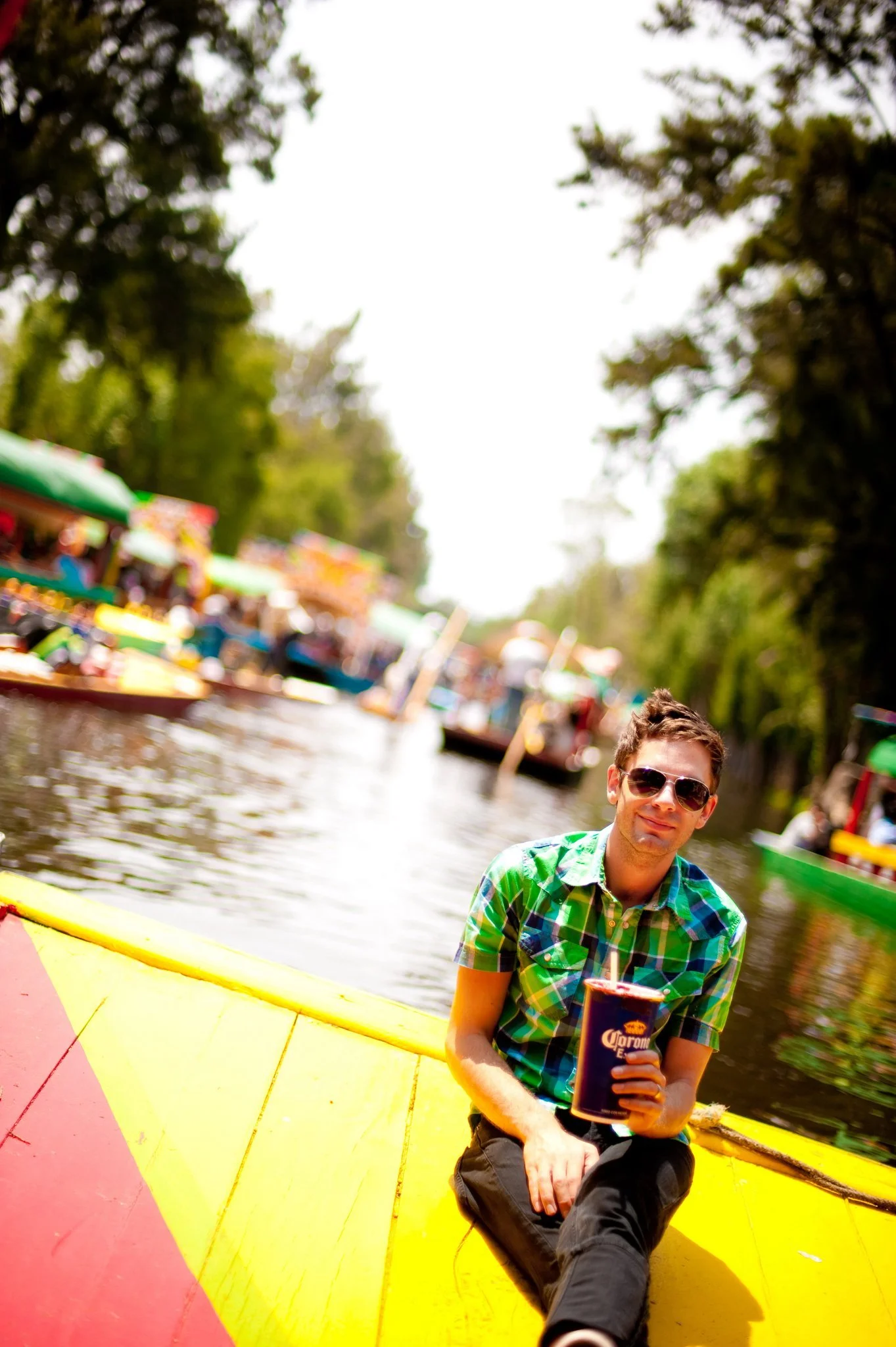 A man with sunglasses and a green plaid shirt sitting on a yellow boat, holding a Corona beer cup, on a river with blurred colorful boats and green trees in the background.