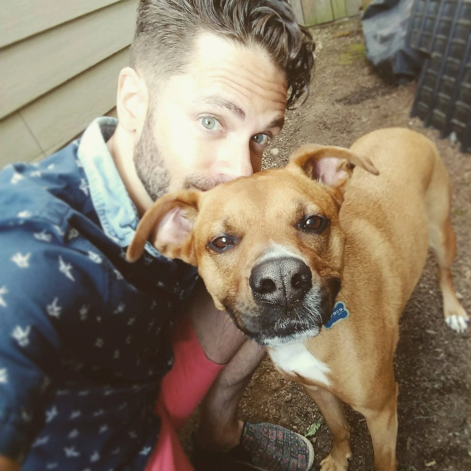 A young man with light skin, curly dark hair, and light-colored eyes is lying on the ground outdoors, next to a large tan and white dog. The man is wearing a dark jacket with white stars and a red shirt, and he is hugging the dog. The dog has a black nose, expressive eyes, and a collar with a blue tag, and it is lying close to the man. The background shows dirt, a wooden fence, and some black objects.