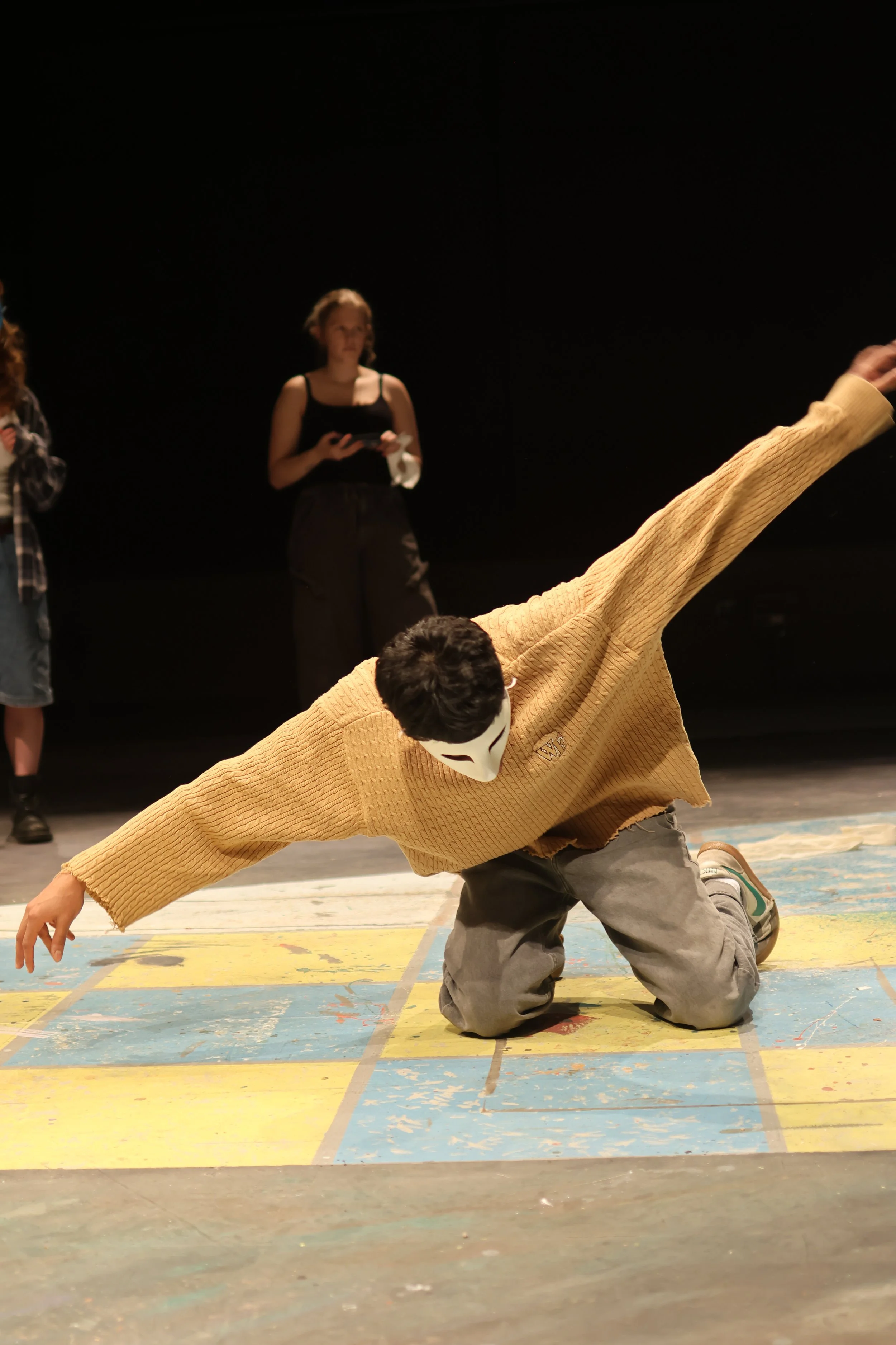 A young man wearing a mask and yellow sweater is performing a dance move on a colorful stage with a black background. Two people are watching in the background, one holding a phone, and another with a hand raised.