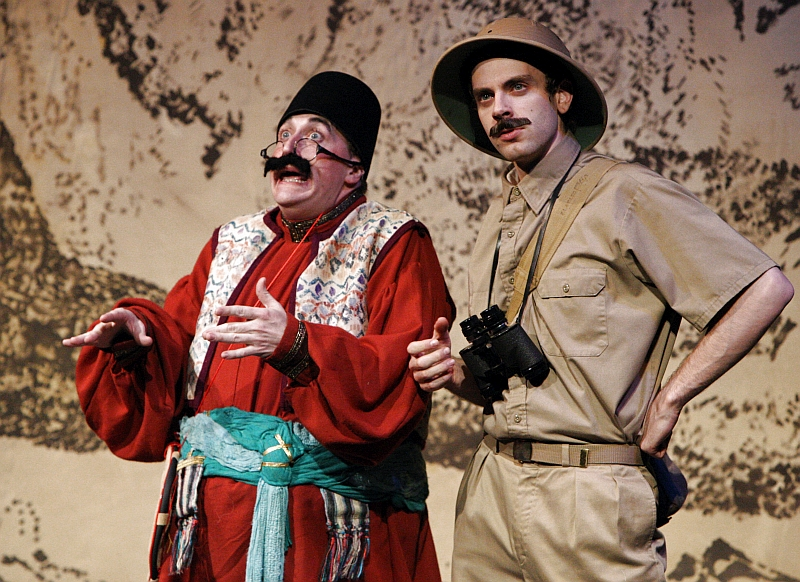 Two actors on stage in costume with a painted backdrop, one dressed as a traditional character with a mustache, glasses, and colorful robes, and the other as an explorer with a khaki shirt, hat, and binoculars around his neck.