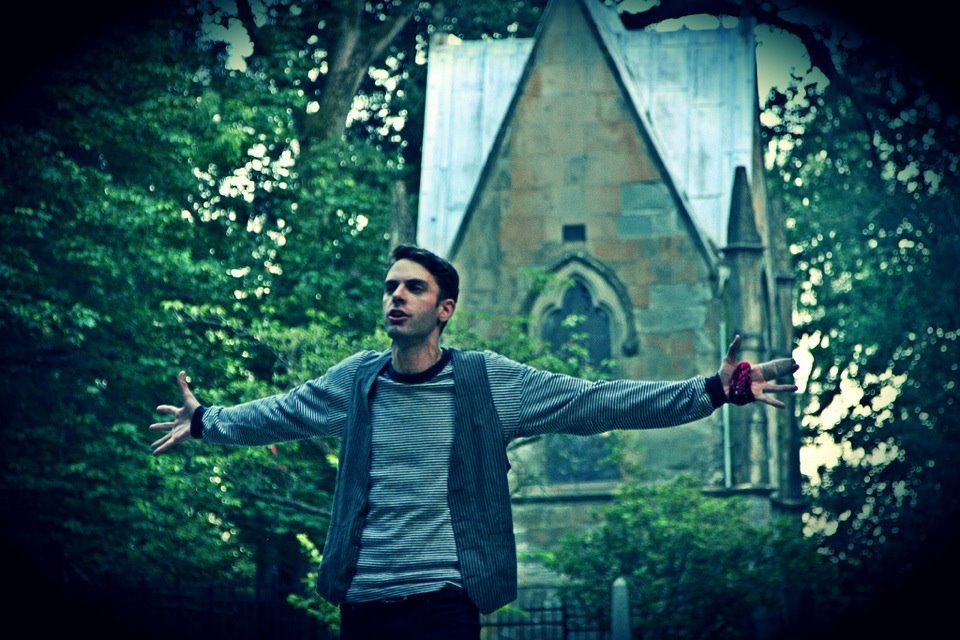 A man with arms outstretched standing in front of a church with Gothic architecture surrounded by trees.