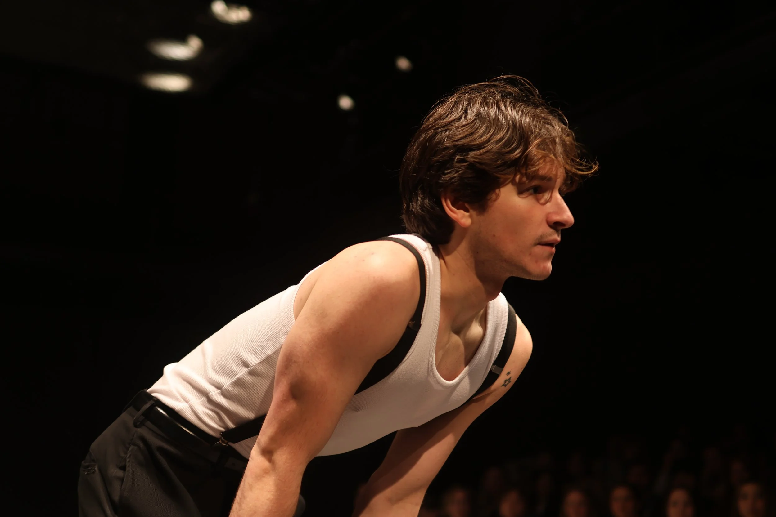 Young man in a white tank top and suspenders bending forward with a dark background.