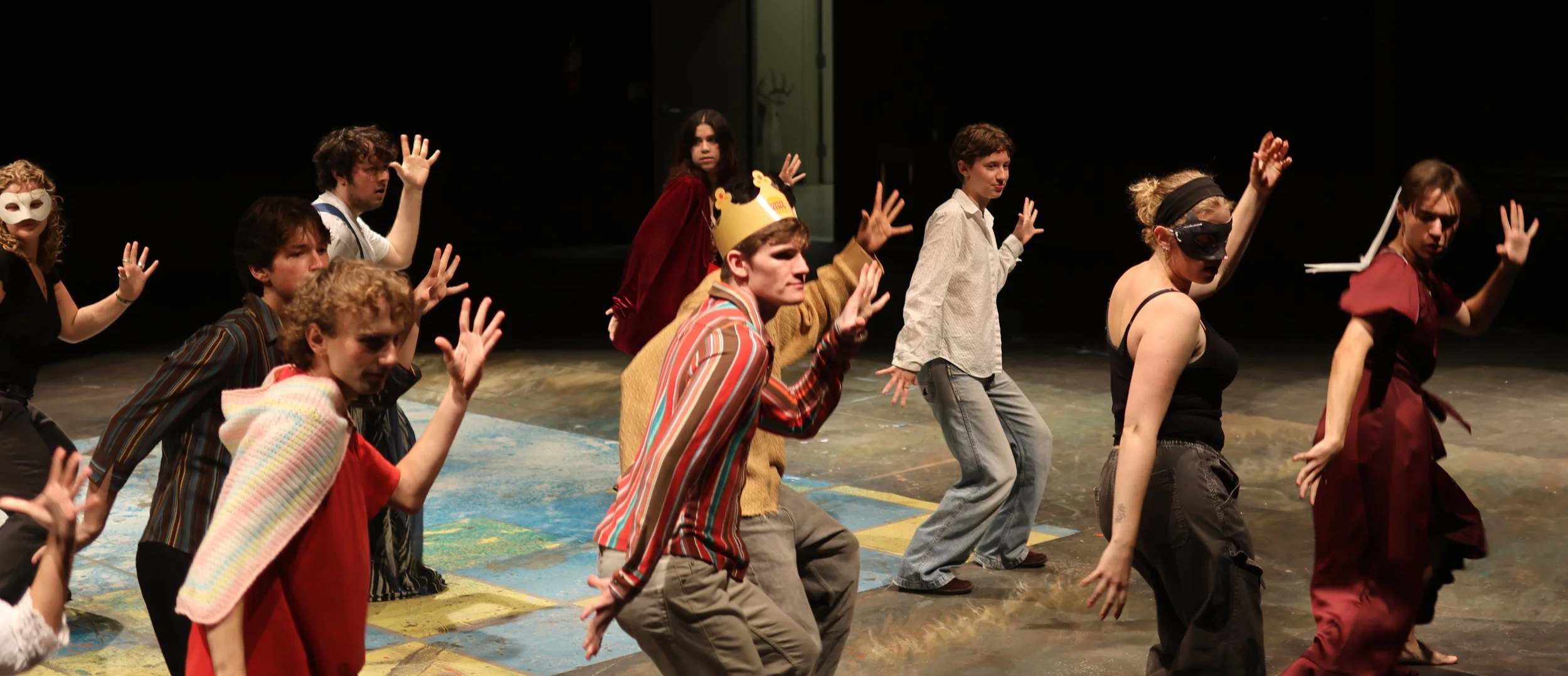Theater actors on stage, some wearing costumes and masks, another in a Burger King paper crown, performing a dance.