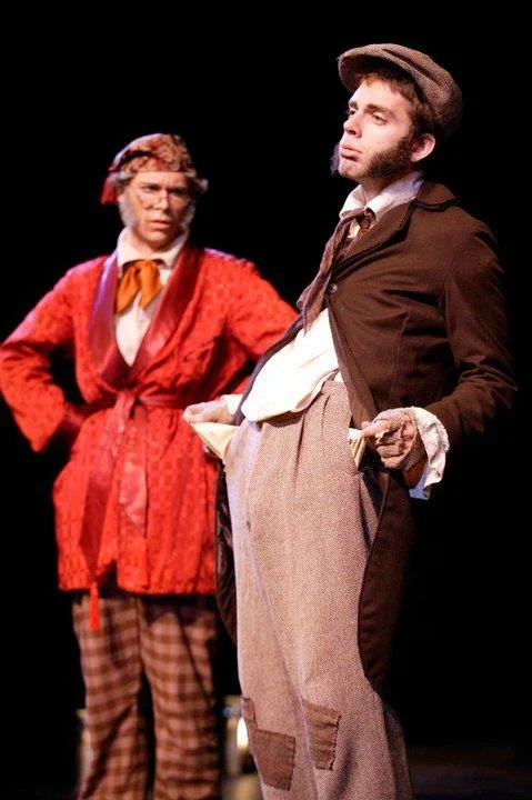 Two actors in period costumes performing on stage; the woman is wearing a red jacket, orange scarf, and plaid pants, while the man is in a brown coat, gray hat, and baggy pants, both with expressive poses.