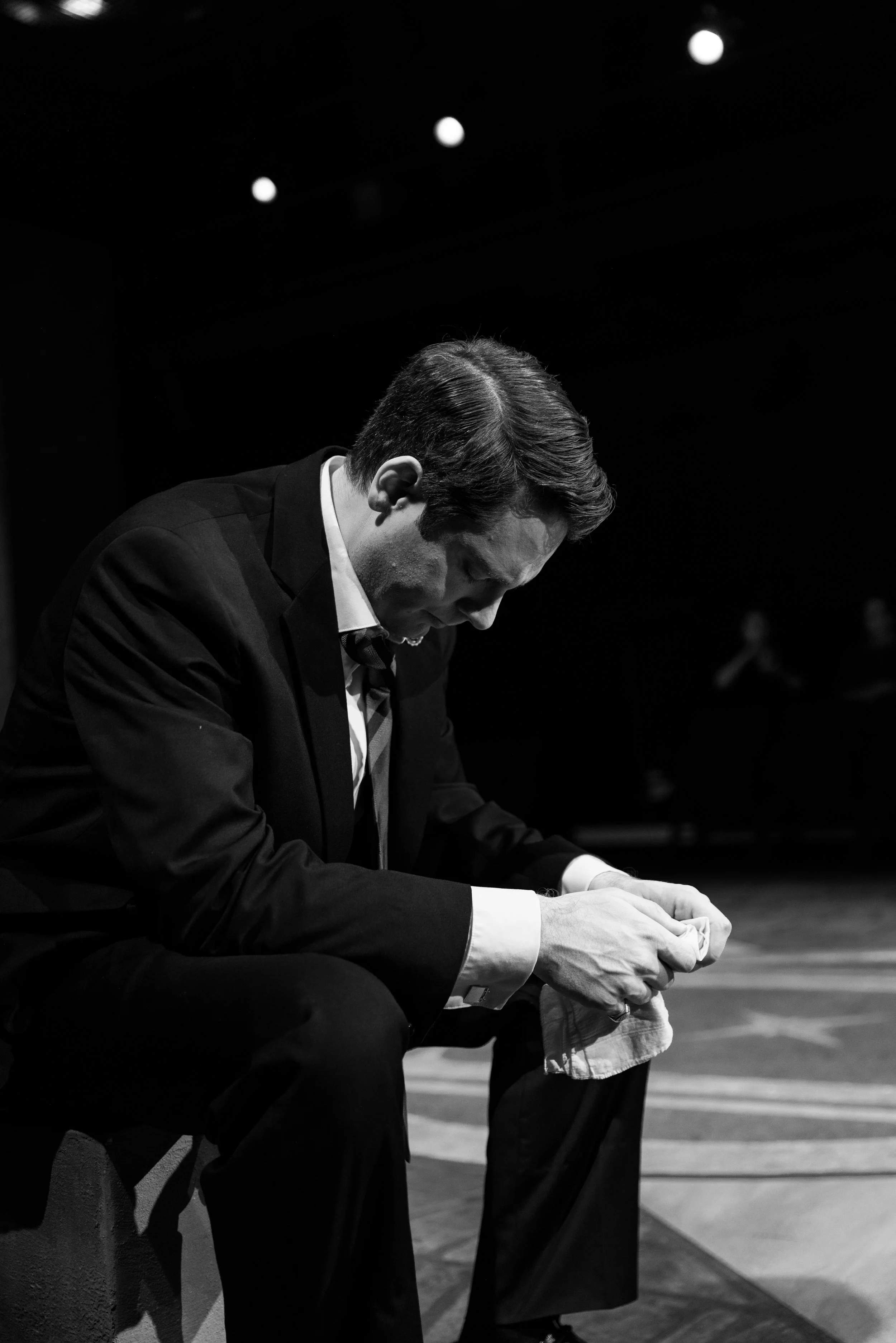 A black and white photo of a man in a suit sitting on a bench, looking down and holding a cloth in his hands, with a dark background and three bright lights overhead.